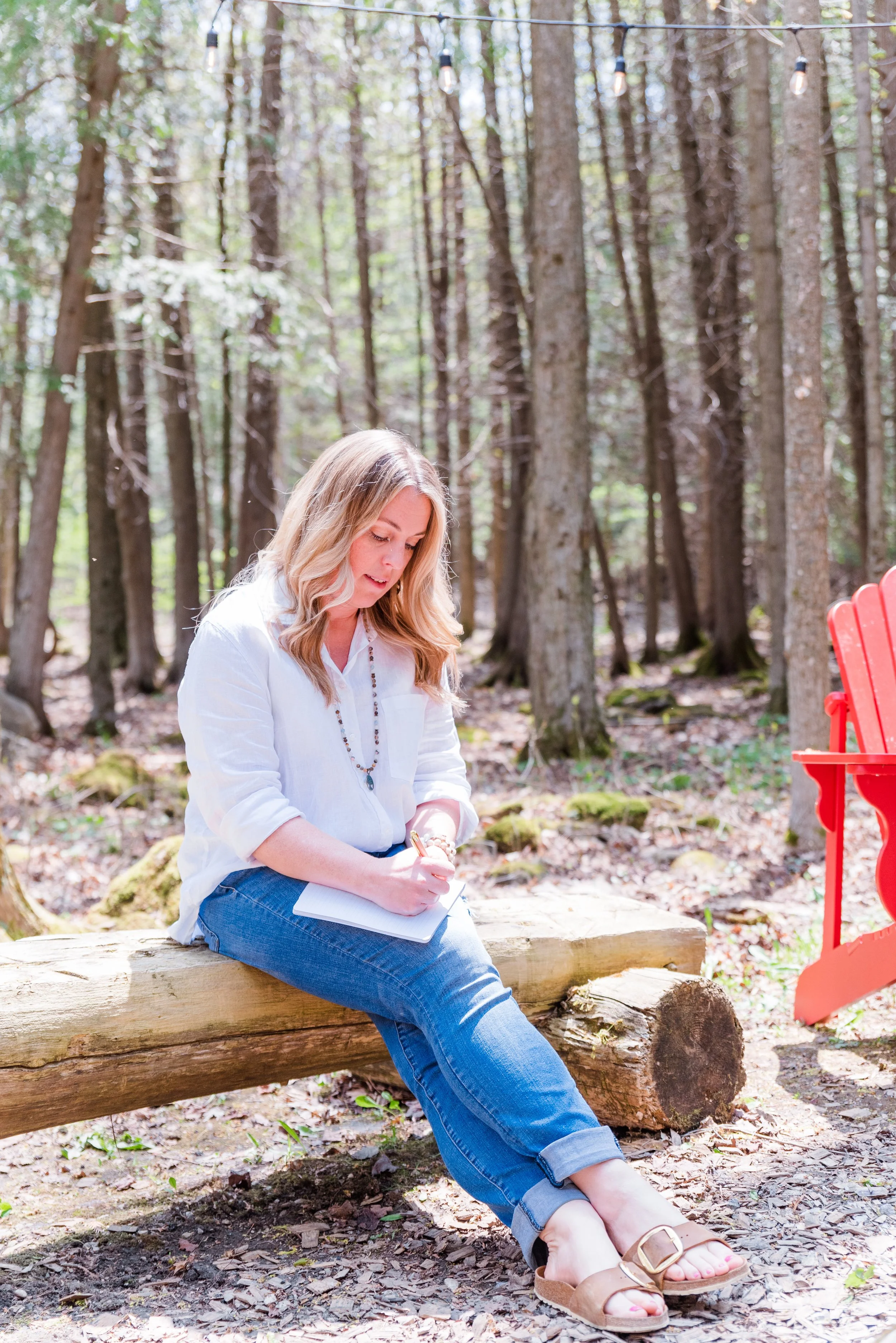 Personal branding photograph of a woman business owner sitting on a log in a forest, writing in a notebook, wearing a white shirt and jeans.