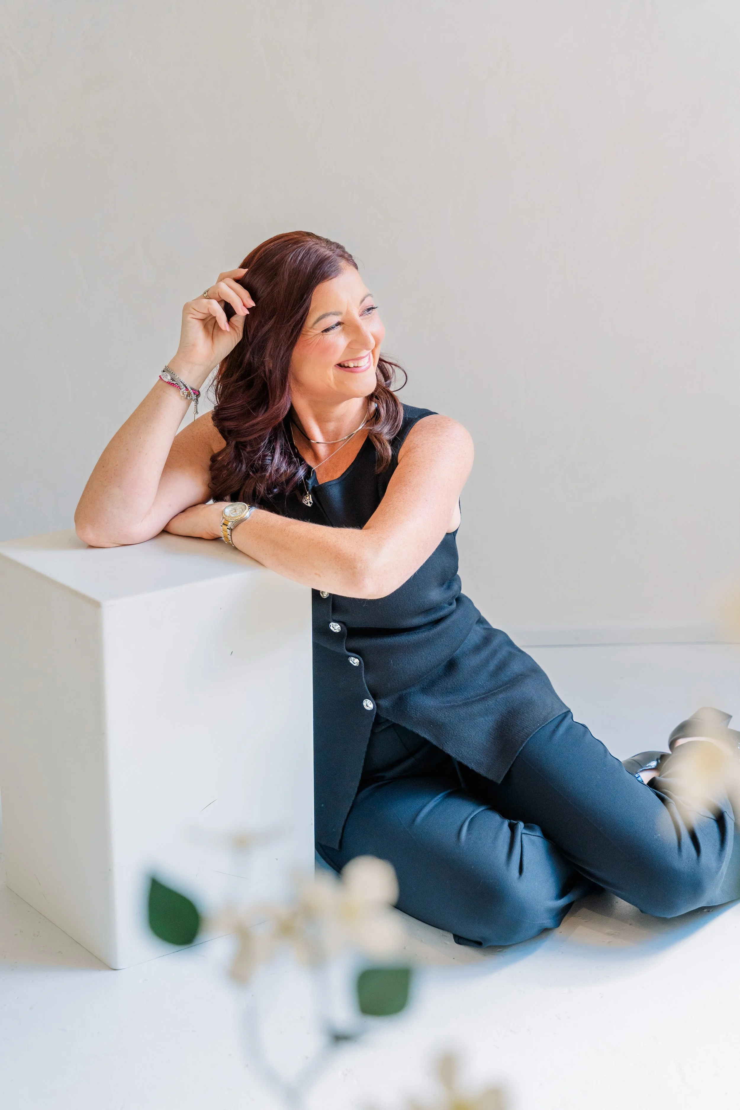 Personal brand photograph of a woman sitting down,  leaning against a white box, smiling and looking off into the distance.