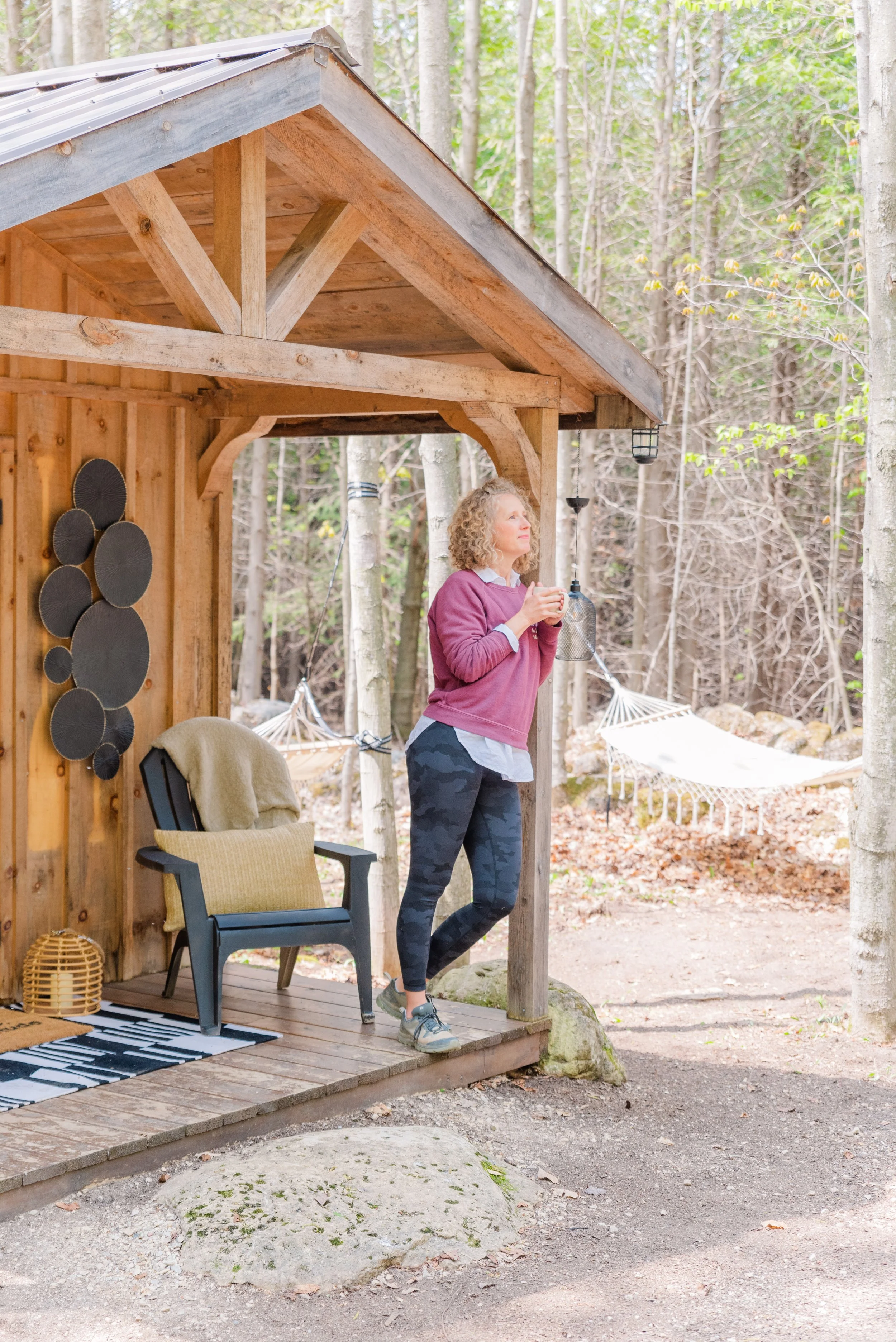 A woman stands on a wooden cabin porch, holding a cup, surrounded by a forest with a hammock in the background. She wears a pink sweater and leggings, with an Adirondack chair and decorative wall art nearby.