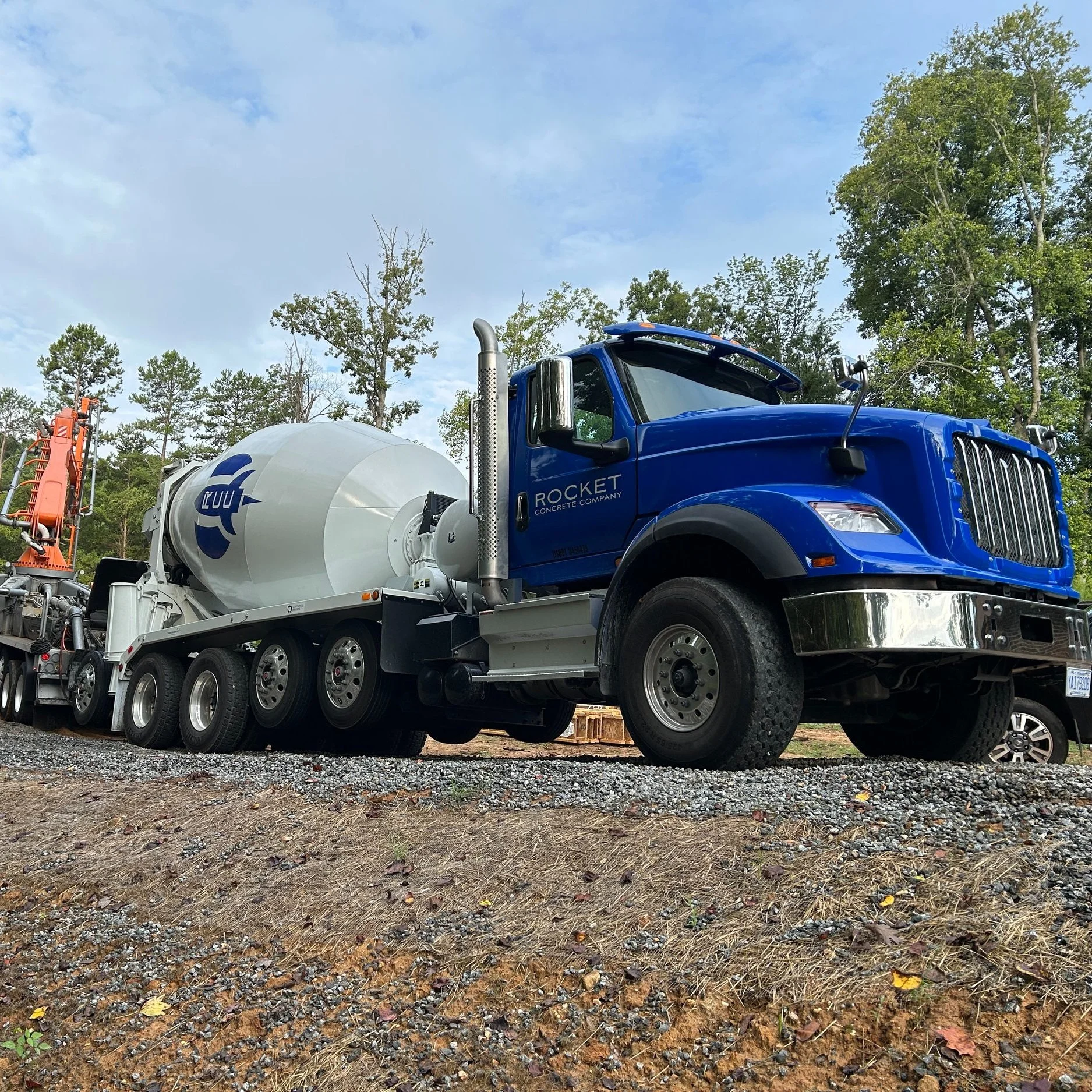 A blue cement mixer truck parked outdoors on a gravel surface with trees and blue sky in the background.