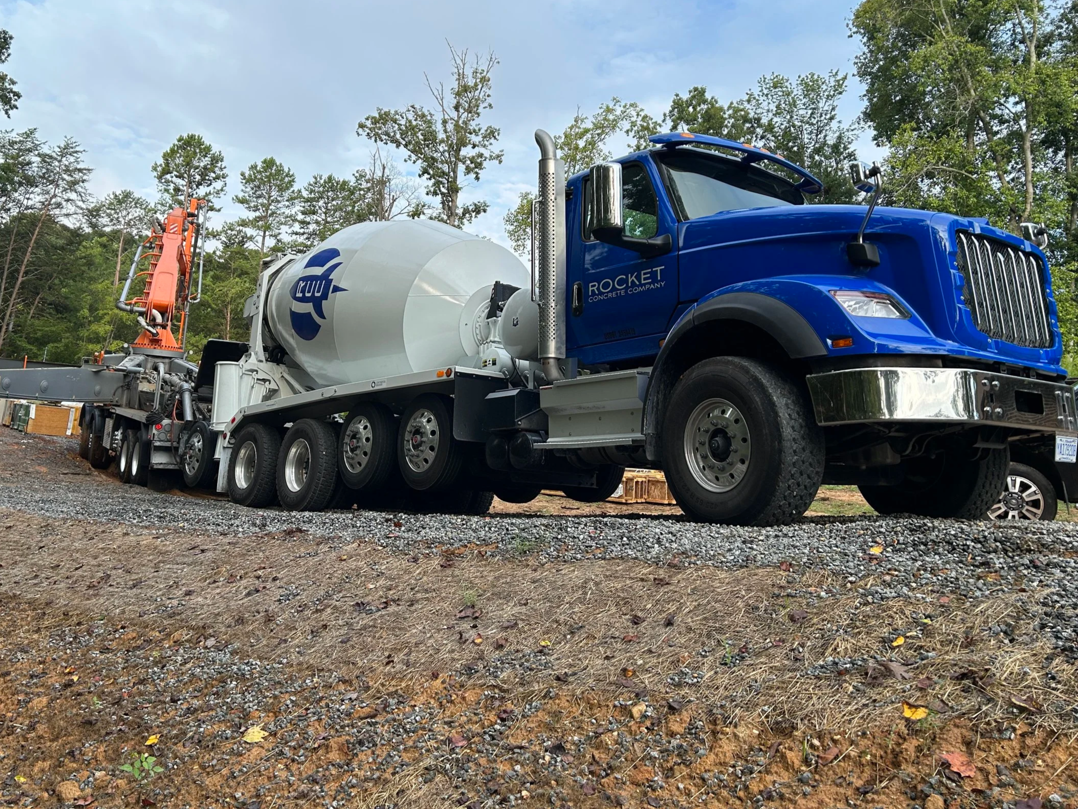 A large blue cement mixer truck with a white mixing drum labeled "RCC" on the side, parked on a construction site in Mooresville NC surrounded by gravel, dirt, and trees in the background.