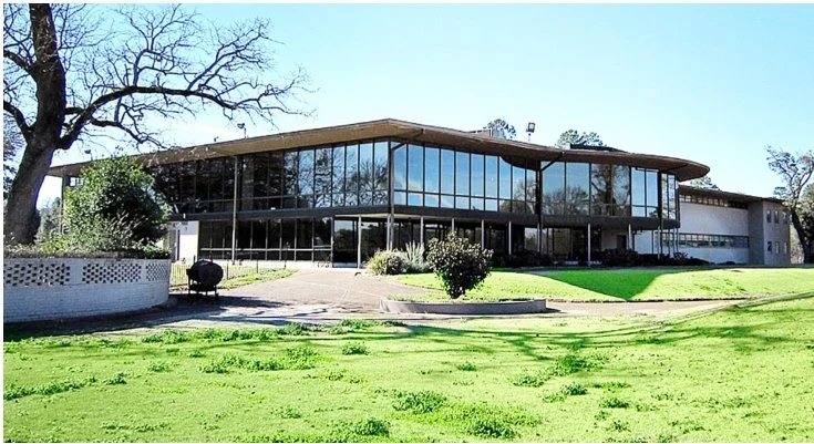 A modern, glass-fronted building with a curved roof, surrounded by a well-maintained lawn and trees.