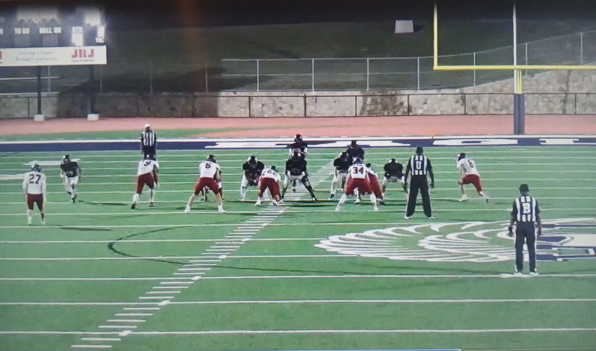 American football game at night showing players lined up at the line of scrimmage on a green field with yard lines and a yellow goalpost in the background.