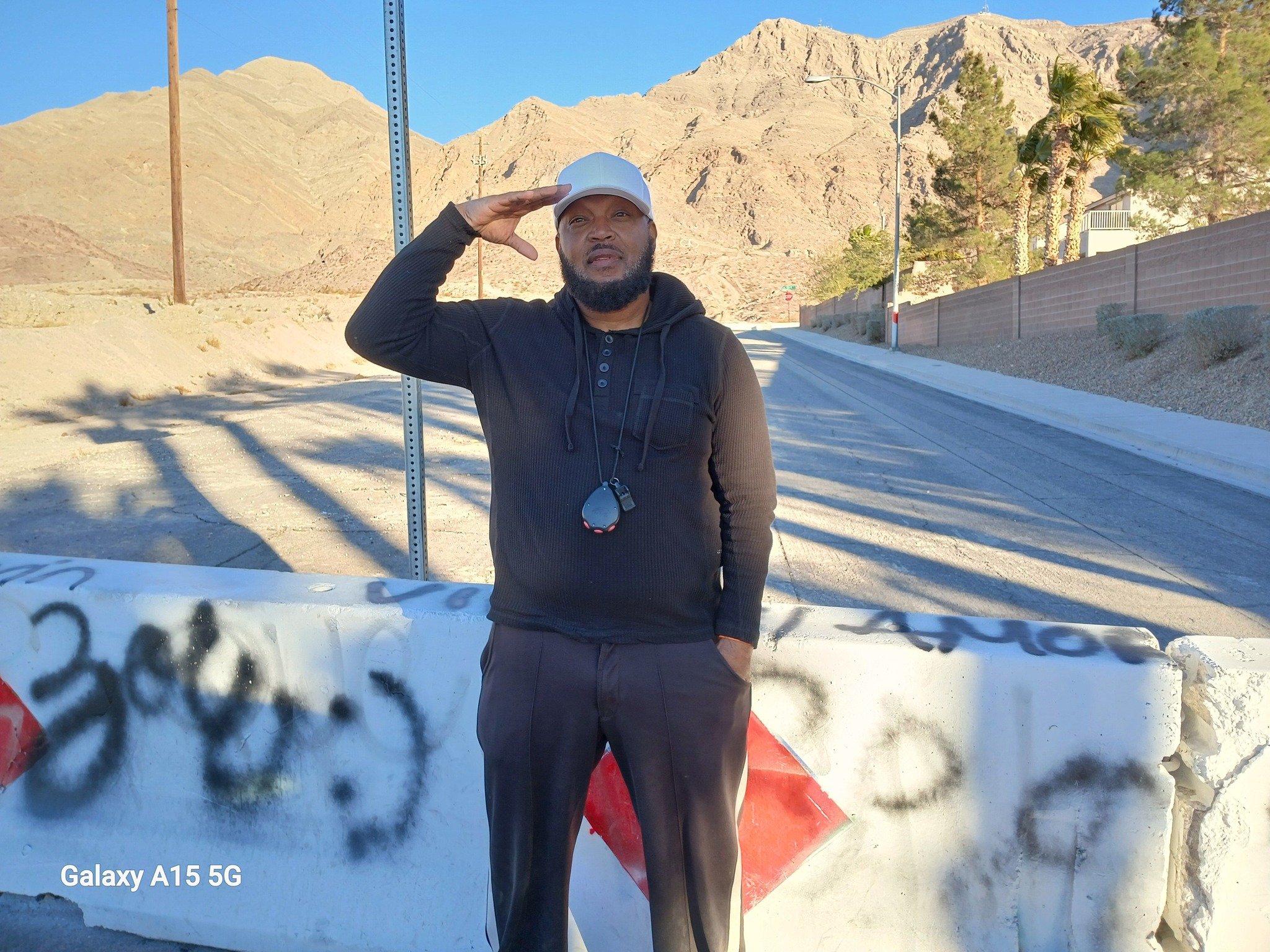 A man saluting while standing outdoors near a concrete barrier with graffiti, mountains in the background, and a clear blue sky.