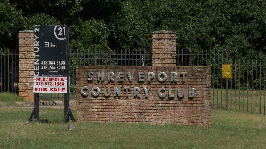 Brick entrance sign for Shreveport Country Club with metal lettering, surrounded by a black metal fence, and an adjacent sign indicating the property is for sale.