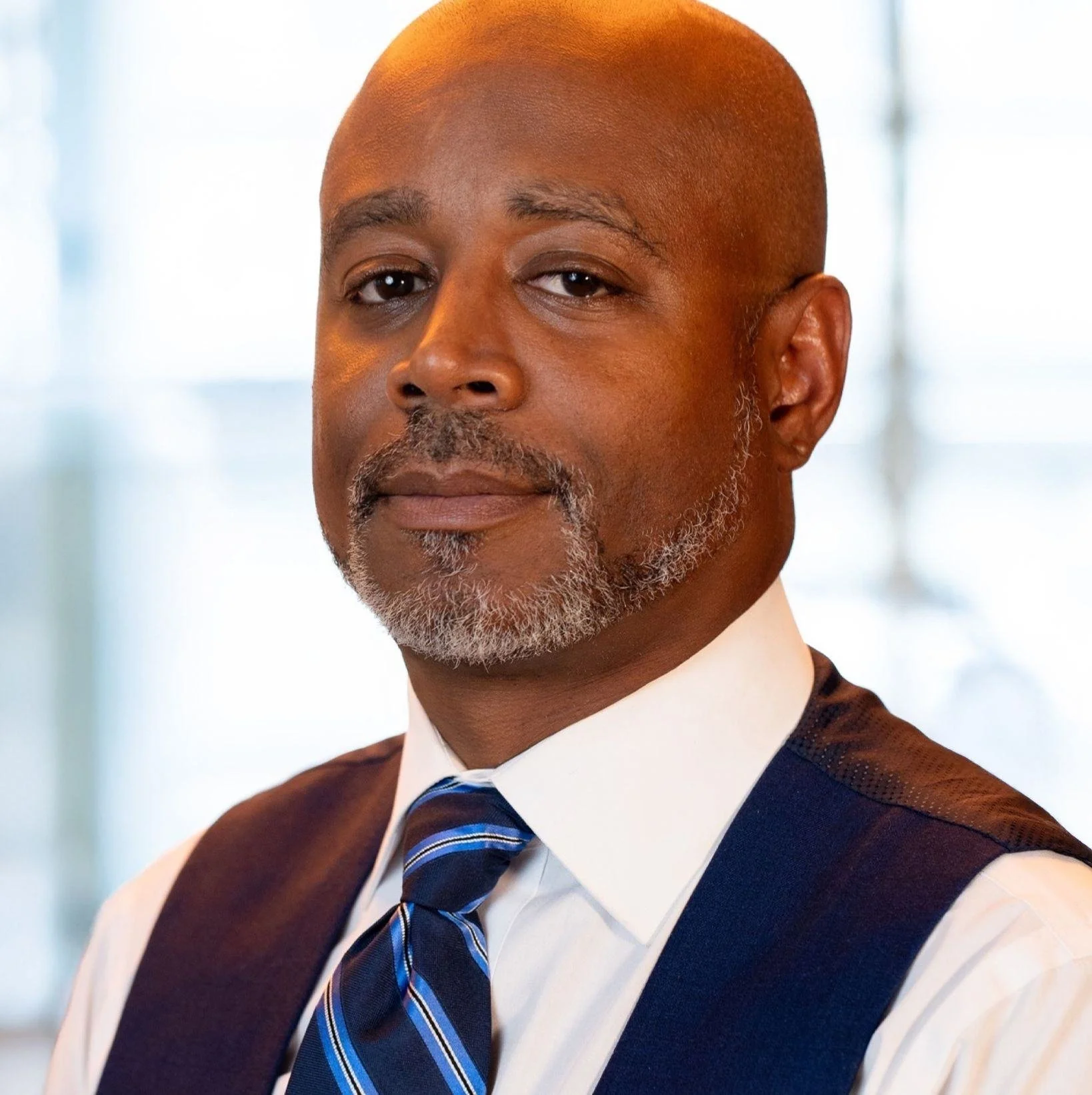 A middle-aged African American man with a gray beard and mustache, dressed in a white shirt, blue striped tie, and dark vest, looking confidently at the camera with a blurred indoor background.