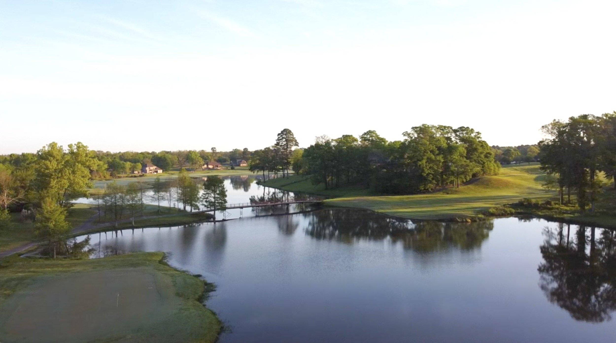 Scenic view of a golf course with water hazards, green grass, and numerous trees under a clear sky.