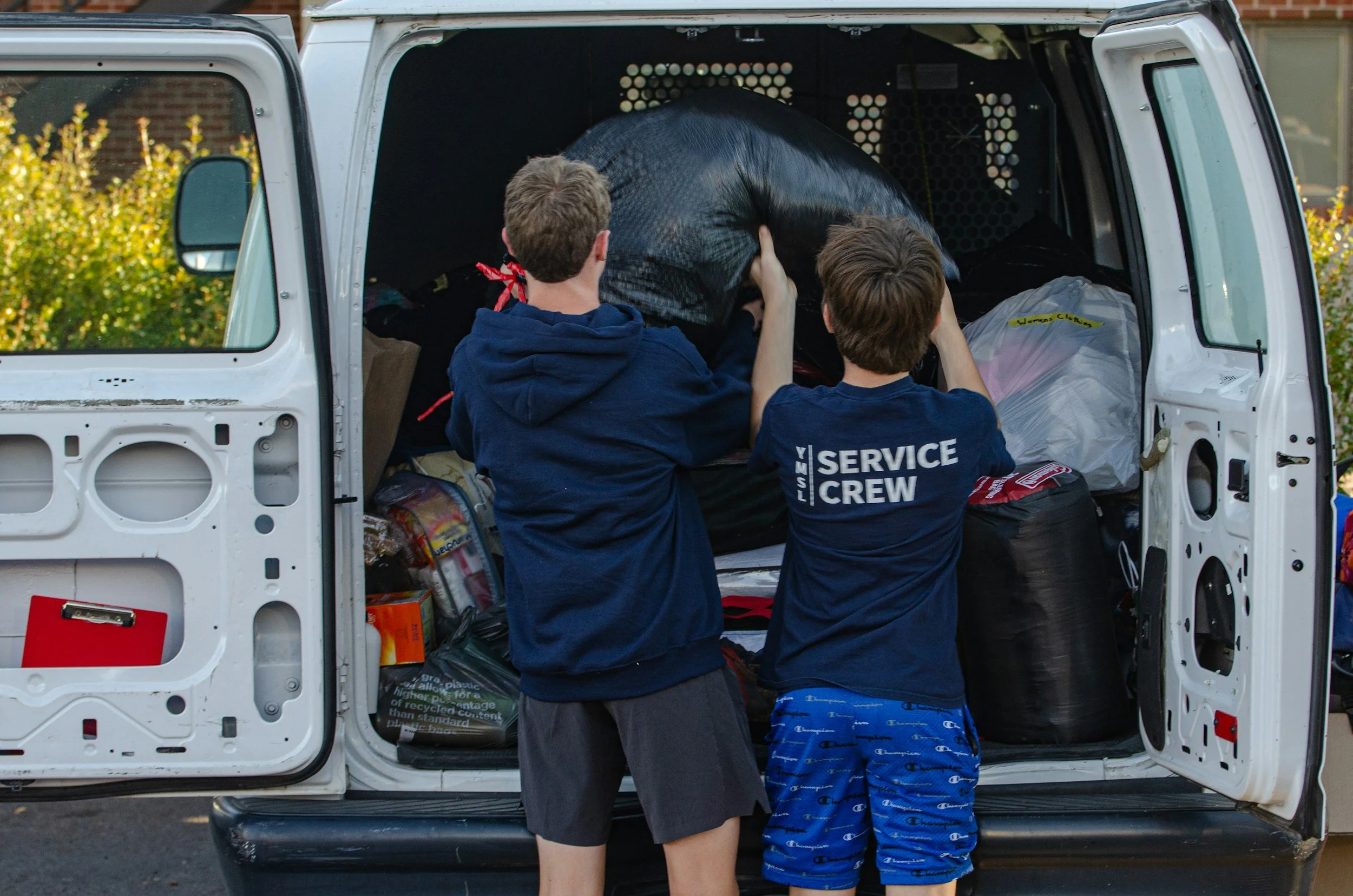 Two boys are loading items into the back of a white van, which is parked outside. One boy is wearing a navy hoodie with service crew printed on the back, and the other boy is wearing blue shorts with Champion logo.