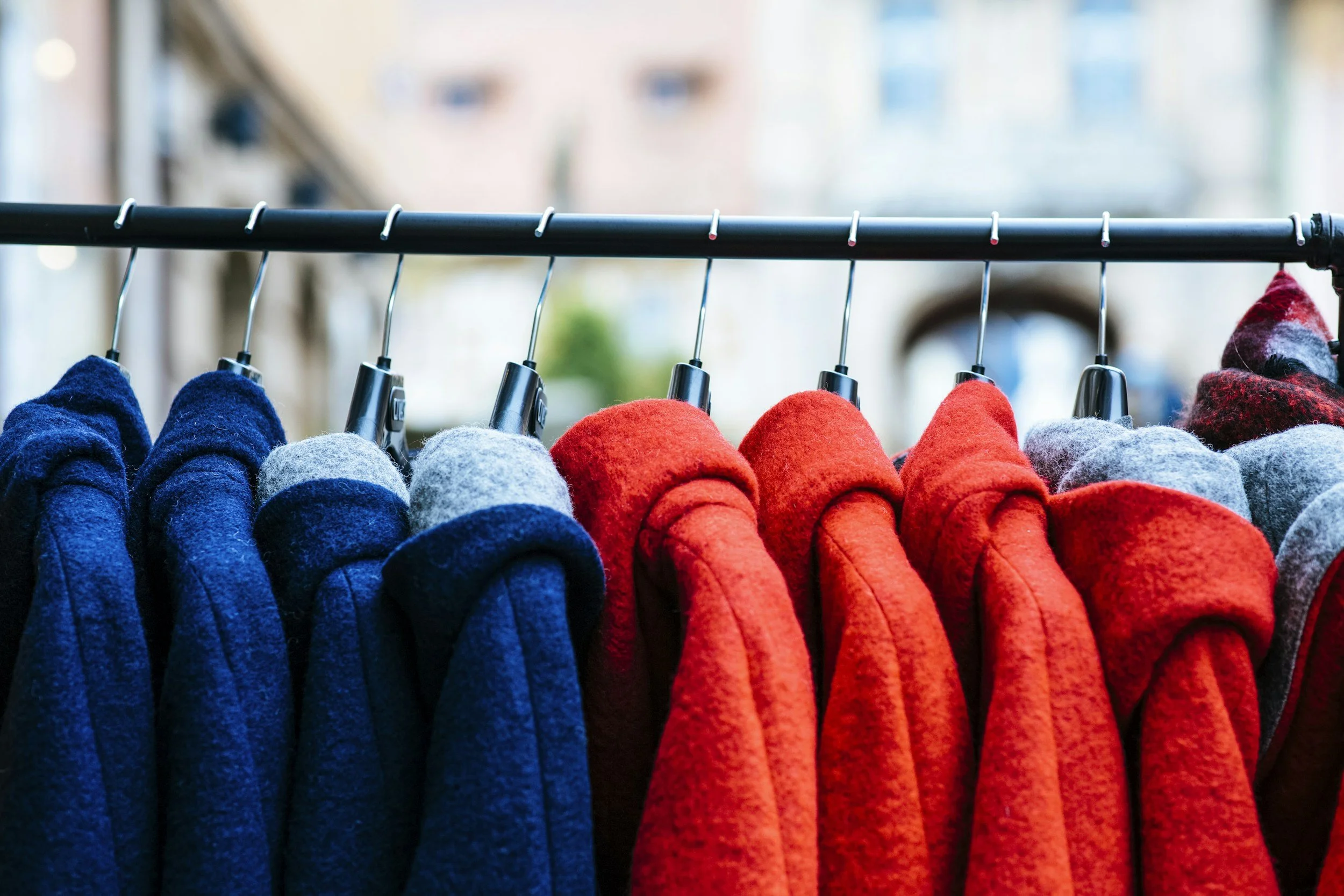 Colorful felt hats in blue, gray, red, and patterned red-black, hanging on a black clothing rack outdoors with blurred city background.