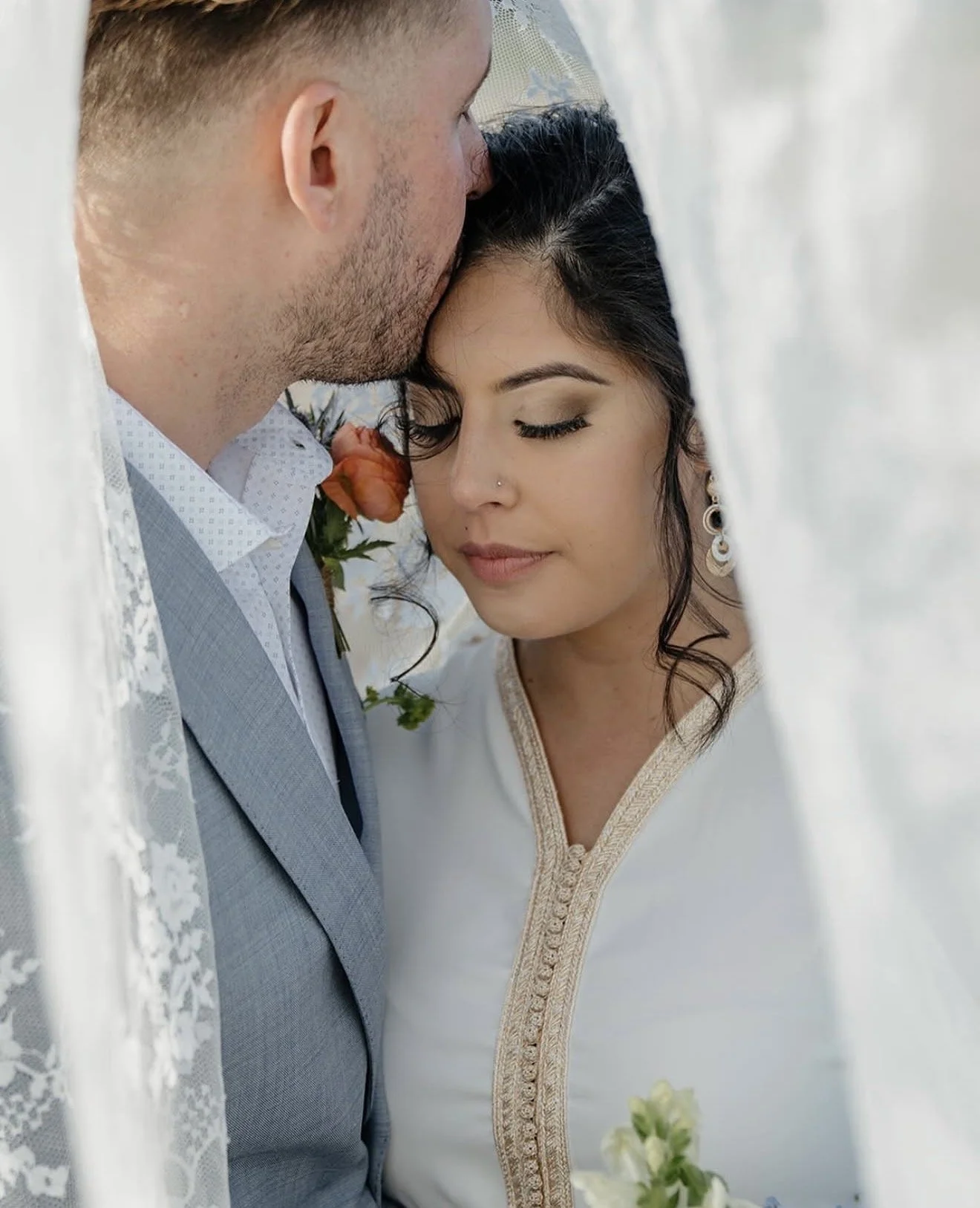 A bride and groom close together under a white veil, with the groom kissing the bride's forehead during their wedding.