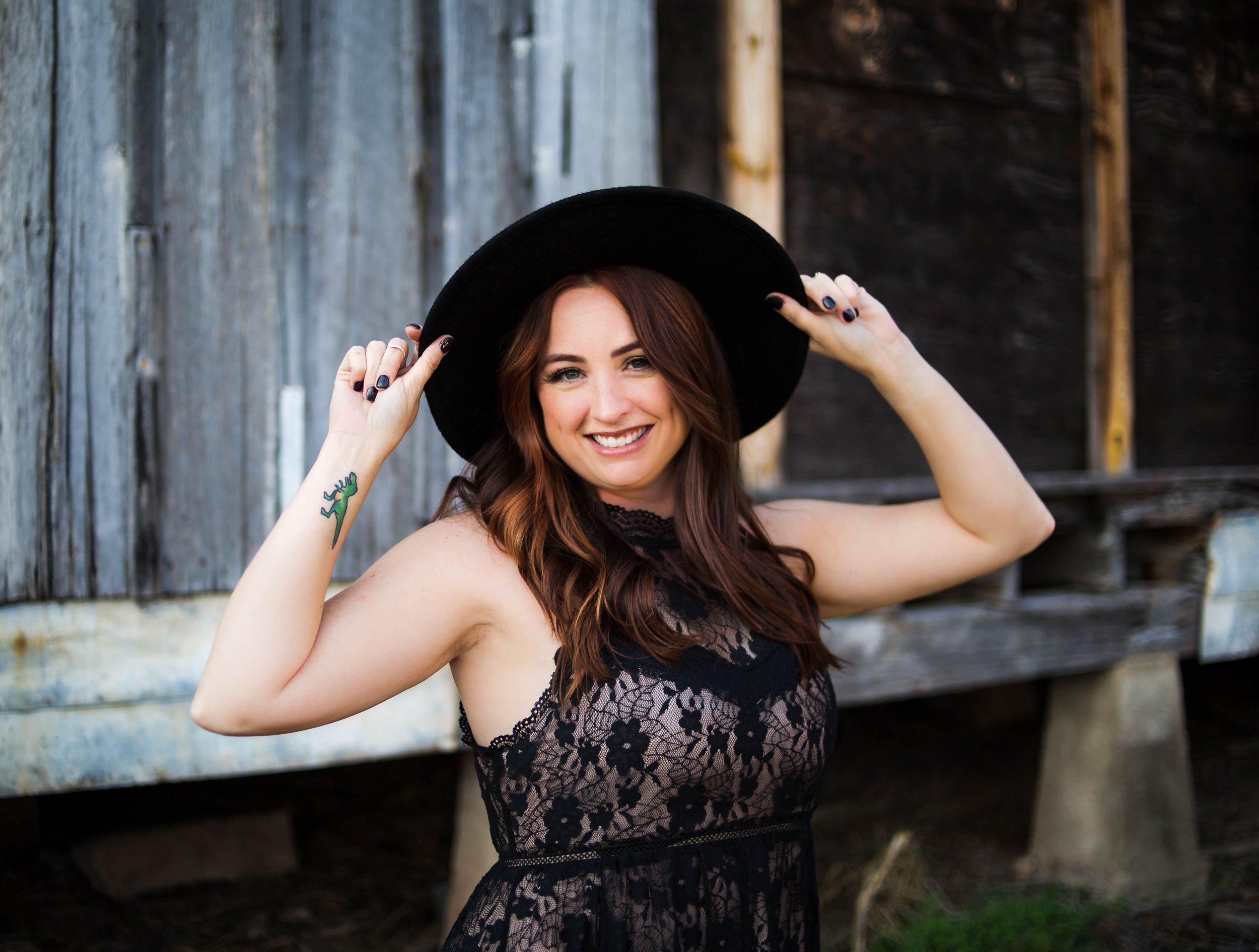 A woman with long red hair and a tattoo of a green lizard on her arm, smiling and wearing a black lace dress and a black hat, standing outdoors in front of a rustic wooden wall.
