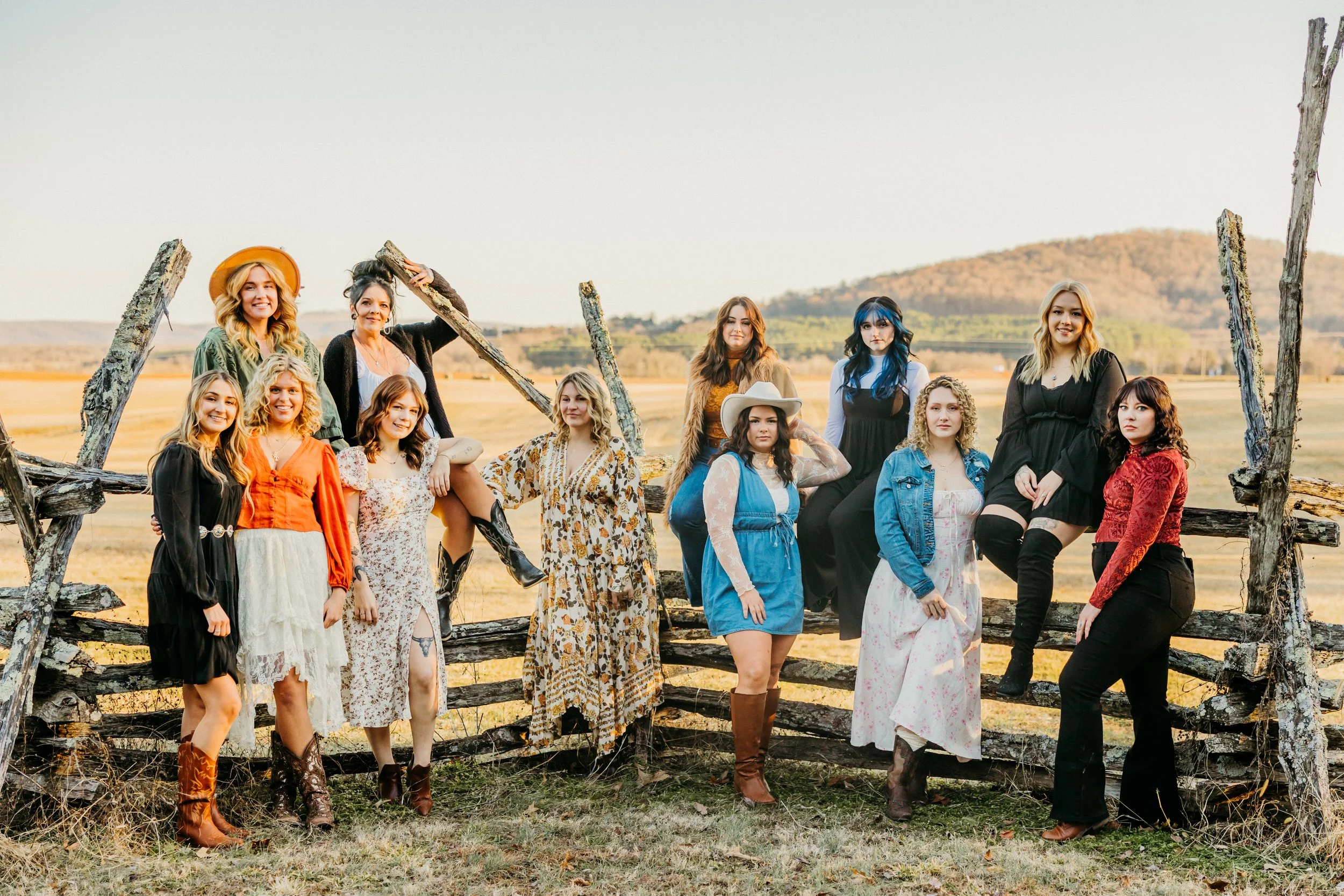 Group of women standing outdoors on a wooden fence in a rural area with open fields and hills in the background, dressed in various colorful and patterned outfits.
