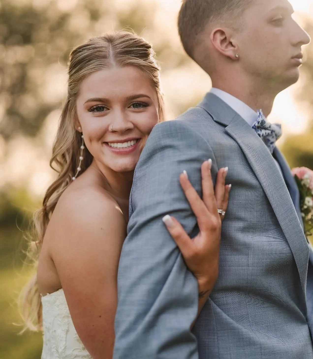 A smiling bride with long blonde hair and a nose ring is hugging a groom from behind, showing her wedding ring. The groom wears a light gray suit, blue shirt, and a patterned bow tie. They are outdoors with a blurred background of trees and warm sunlight.