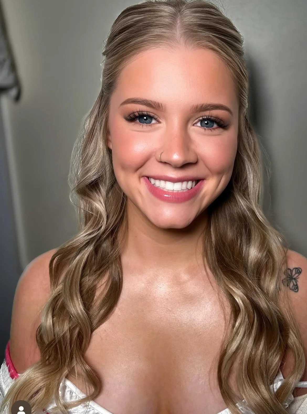 A young woman smiling with long wavy blonde hair, blue eyes, makeup, and a nose ring, wearing a white off-the-shoulder top with a butterfly tattoo on her shoulder.