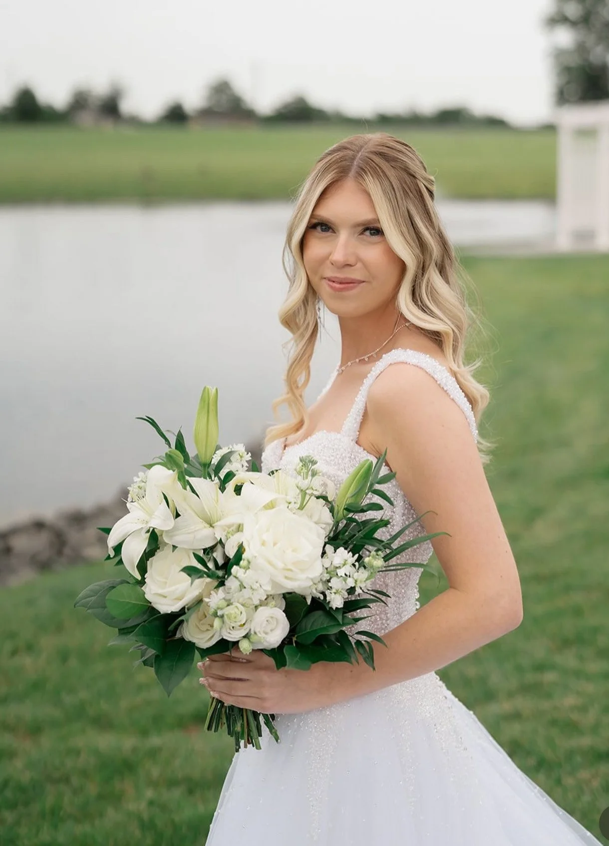 A bride in a white wedding dress holding a large bouquet of white lilies, roses, and greenery, standing outdoors near a pond with blurred green grass and trees in the background.