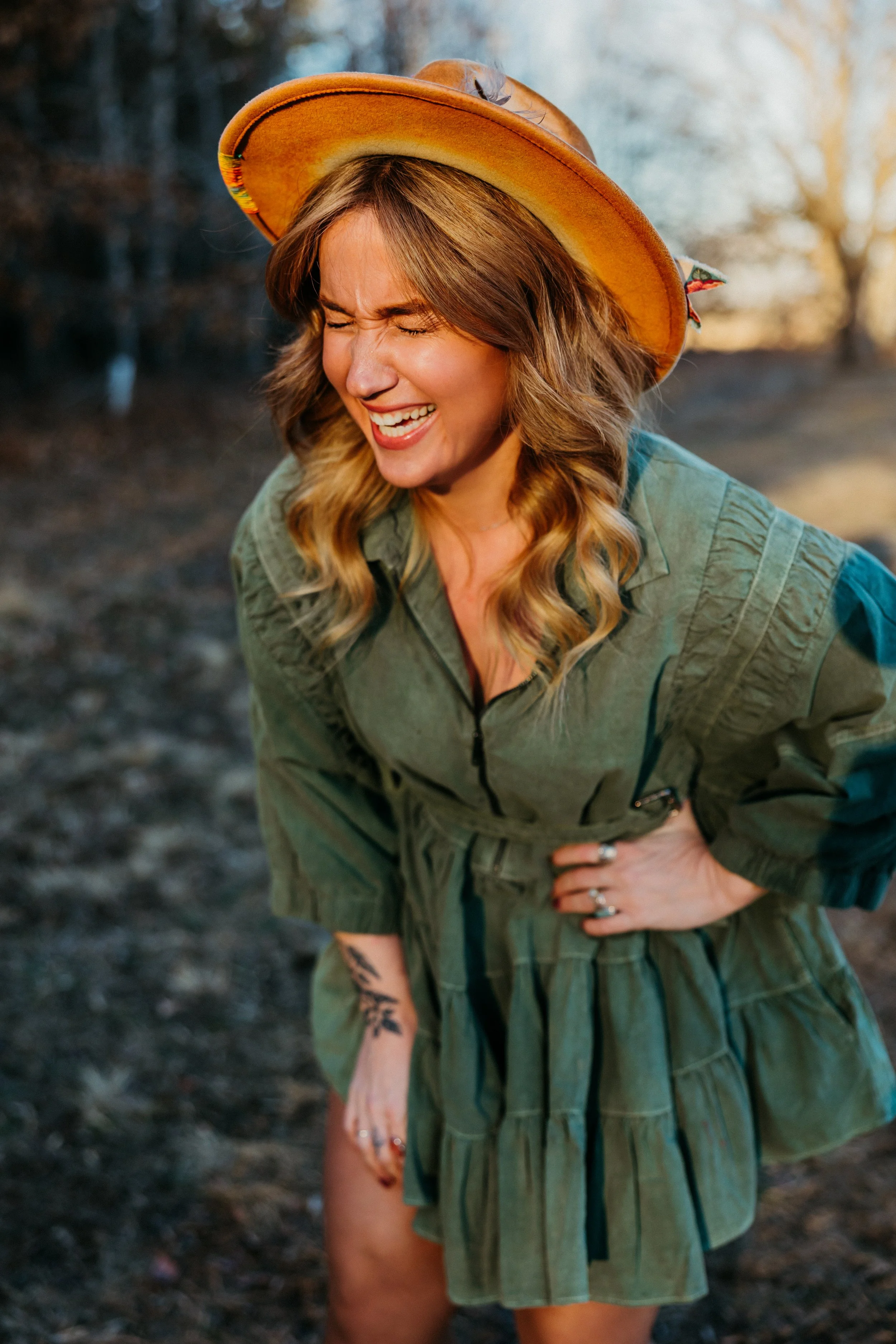 A woman with long blonde hair, wearing a wide-brimmed hat and green outdoor jacket, smiling and laughing outside in a natural setting.