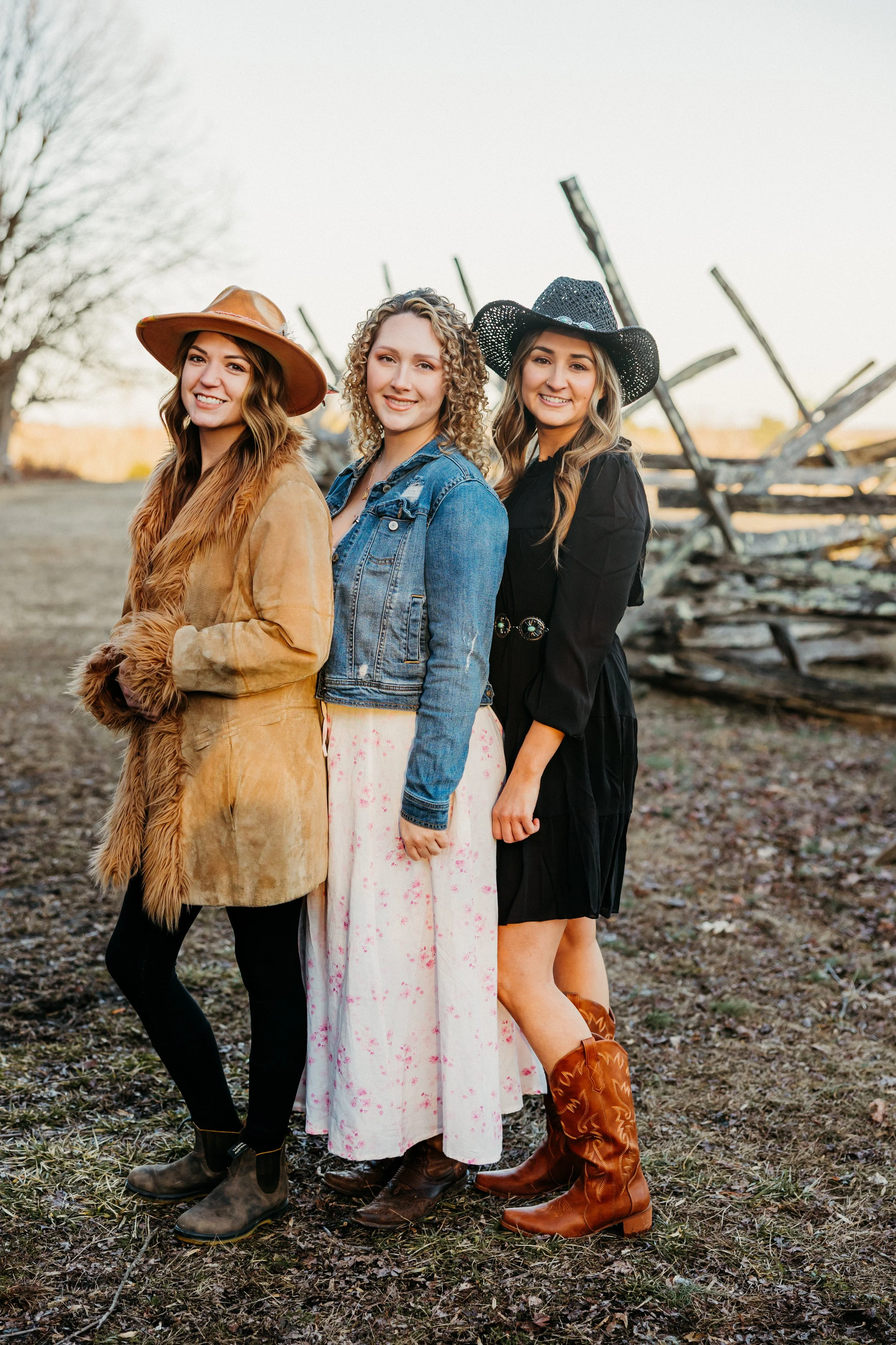 Three women dressed in cowgirl outfits standing outdoors with a rustic wooden fence in the background during daytime.