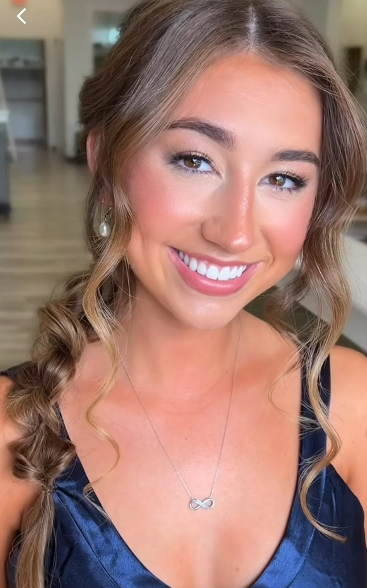 A smiling woman with long, curly hair, wearing pearl earrings and a silver infinity necklace, dressed in a navy blue dress, indoors with a blurred background.