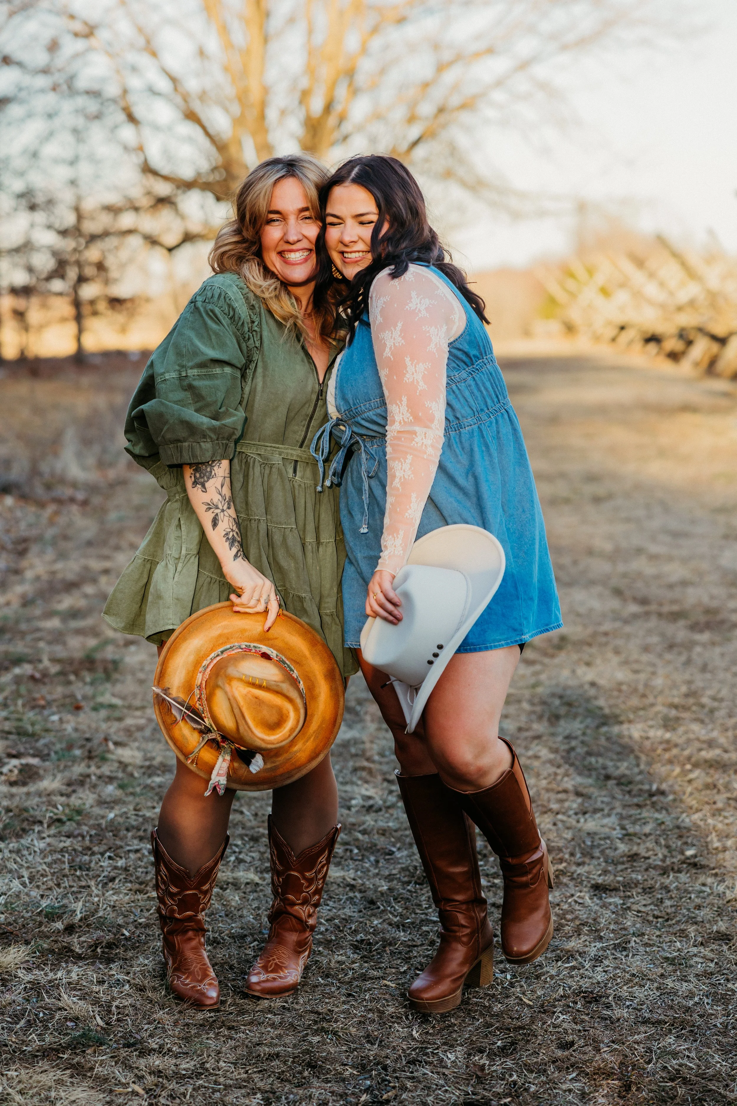 Two women standing outdoors on a grassy area, smiling and laughing together. They are dressed in Western-style clothing, wearing cowboy boots and holding hats and a guitar. The background features trees with some leaves and the scene appears to be during sunset or golden hour.