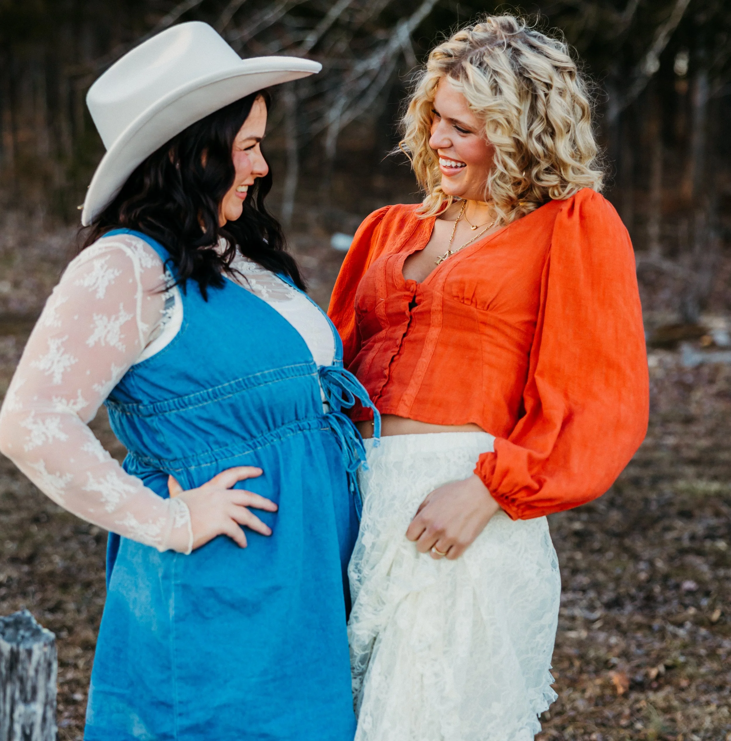 Two women, one with dark hair wearing a white cowboy hat and blue and white lace dress, and the other with blonde curly hair wearing an orange dress, standing close together outdoors and smiling at each other.