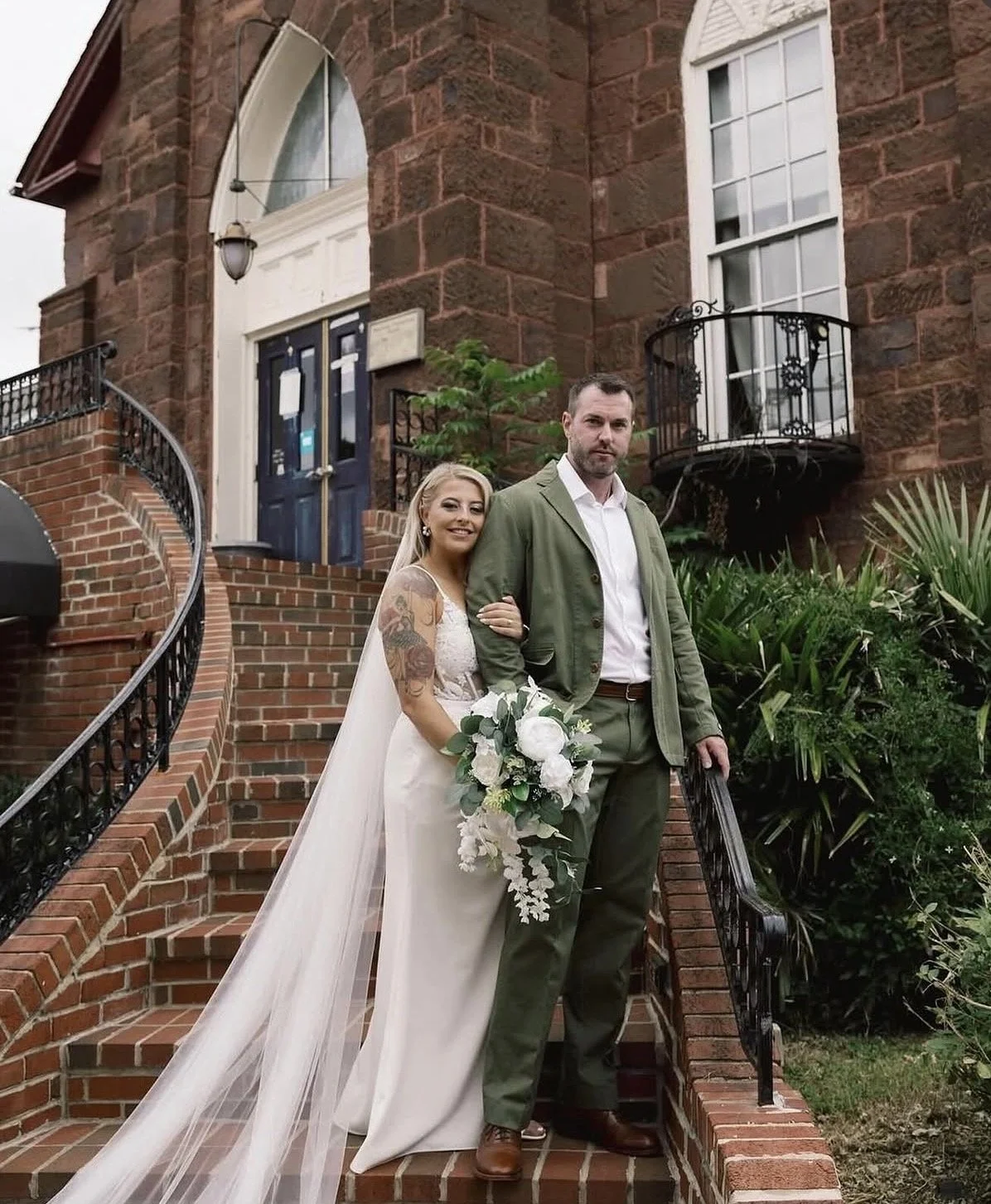 A bride and groom standing on the stairs of a red brick building, with the bride holding a bouquet and smiling, and the groom looking at the camera. The bride has tattoos on her arm and wears a white wedding dress with a long veil. The groom wears a green suit jacket and brown shoes. There are plants and greenery around them.