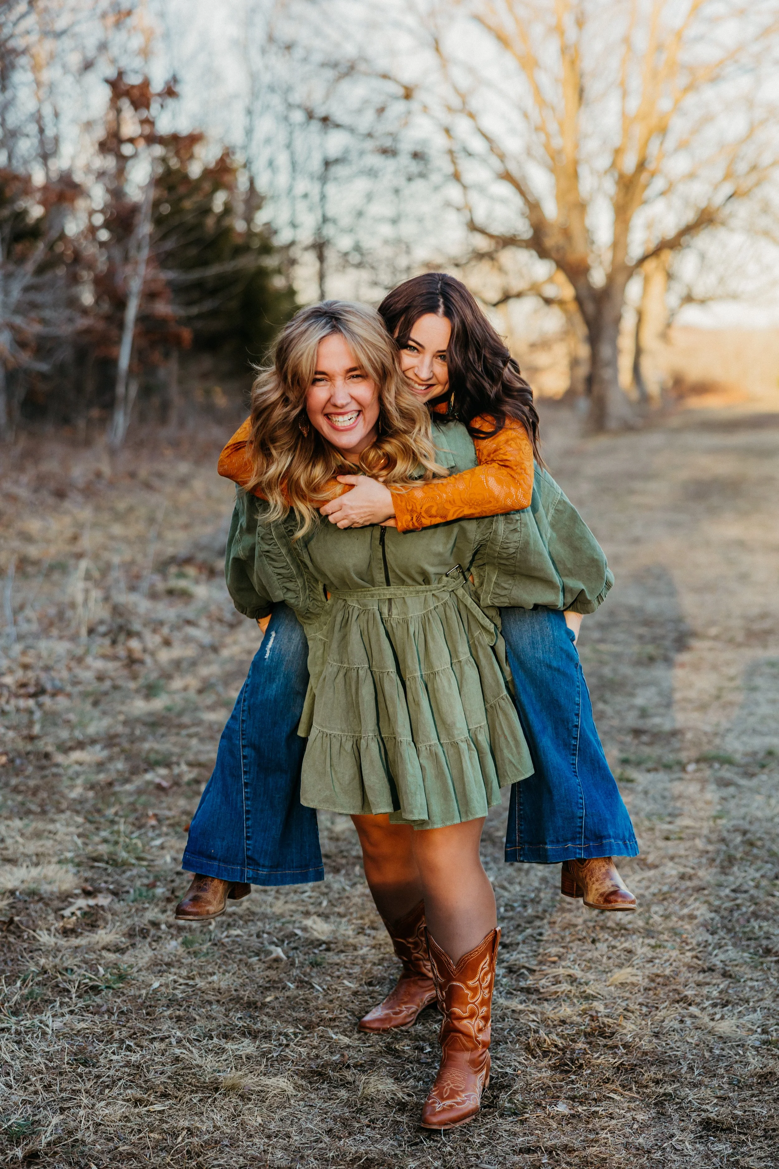 Two women, one with blonde curly hair and one with dark wavy hair, smiling and hugging in an outdoor setting with trees and dry grass, during autumn or winter.