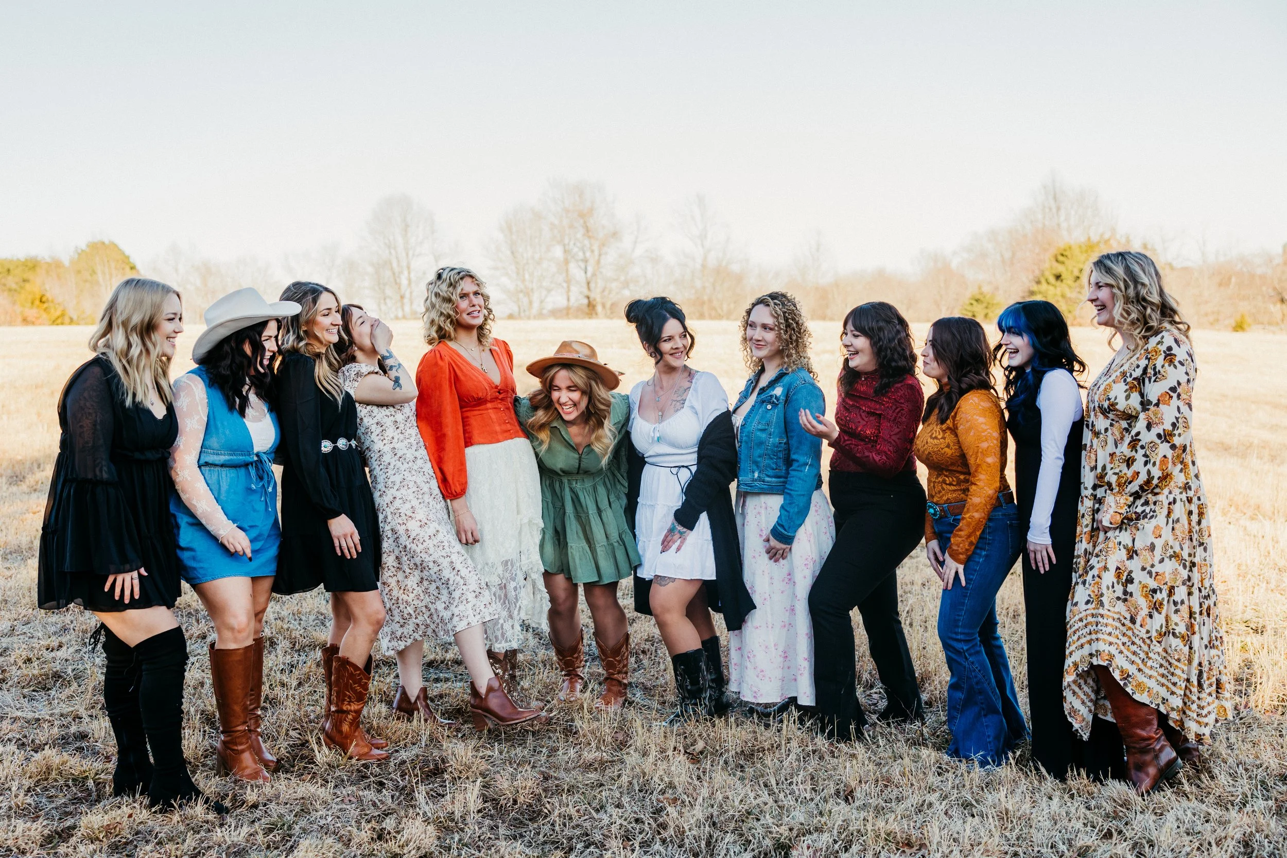 A group of twelve women standing outdoors in a field, laughing and talking to each other during daytime. They are dressed in casual, bohemian-style clothing with various patterns and colors, some wearing hats and boots. The background features a clear sky and distant trees.