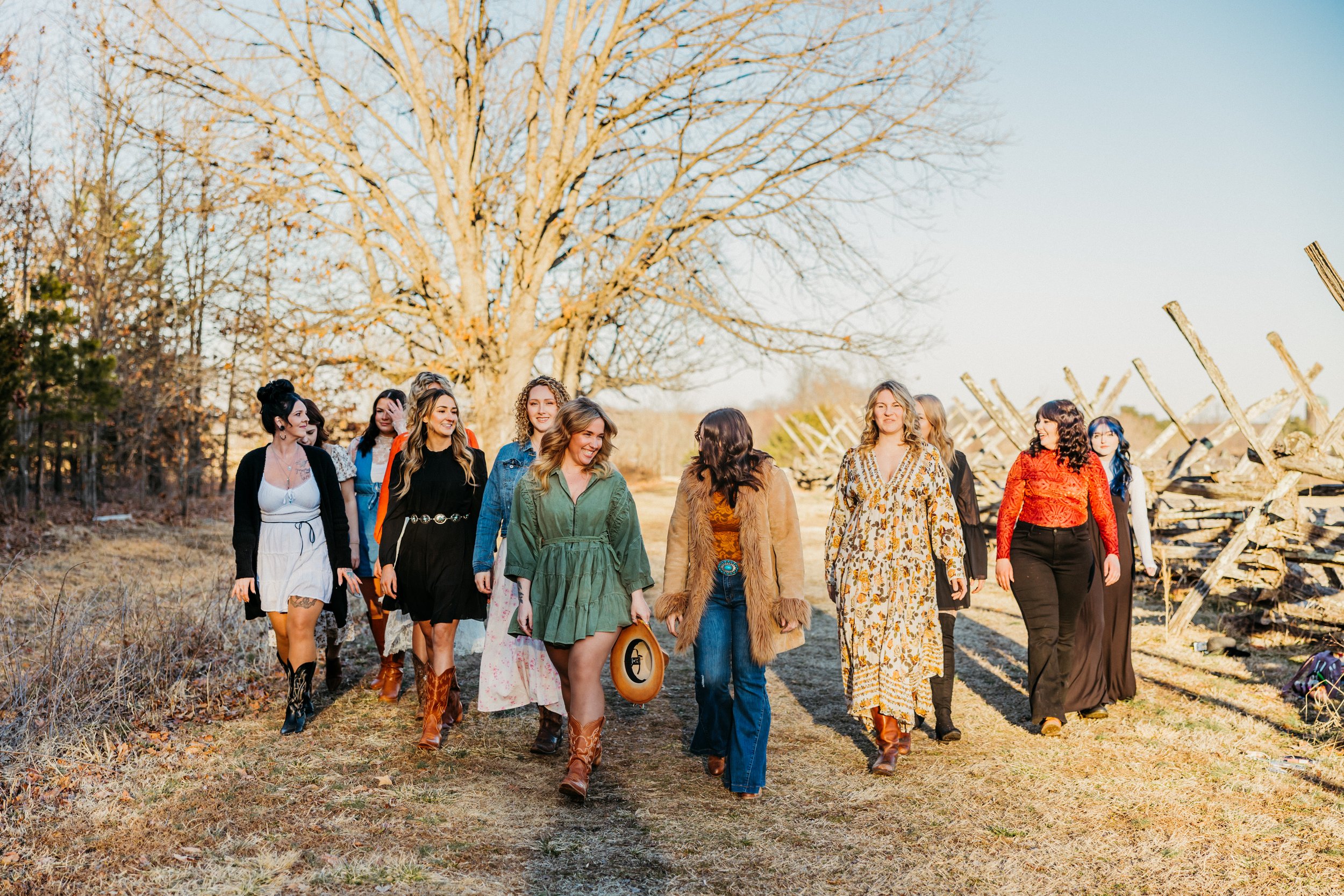 A group of women dressed in casual and bohemian style clothing walking together along a dirt path outdoors in a rural area during late afternoon. The background features a leafless tree and a rustic wooden fence.