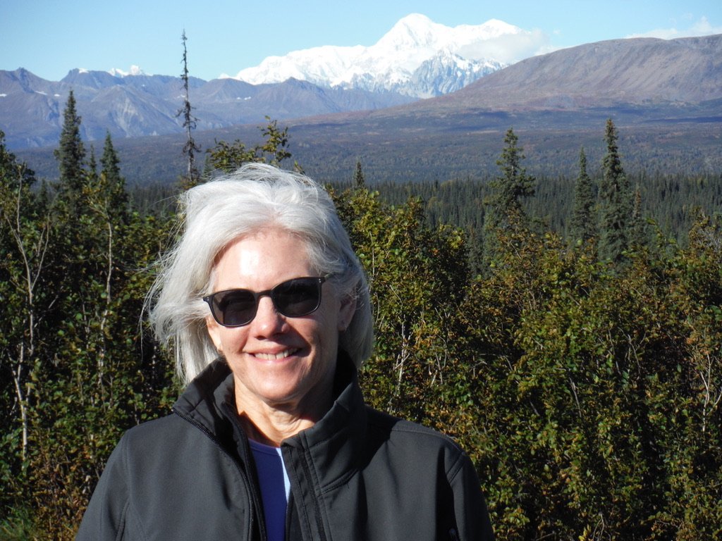 Smiling woman with gray hair wearing sunglasses and a black jacket standing outdoors in a mountainous landscape with snow-capped peaks and dense green forest.