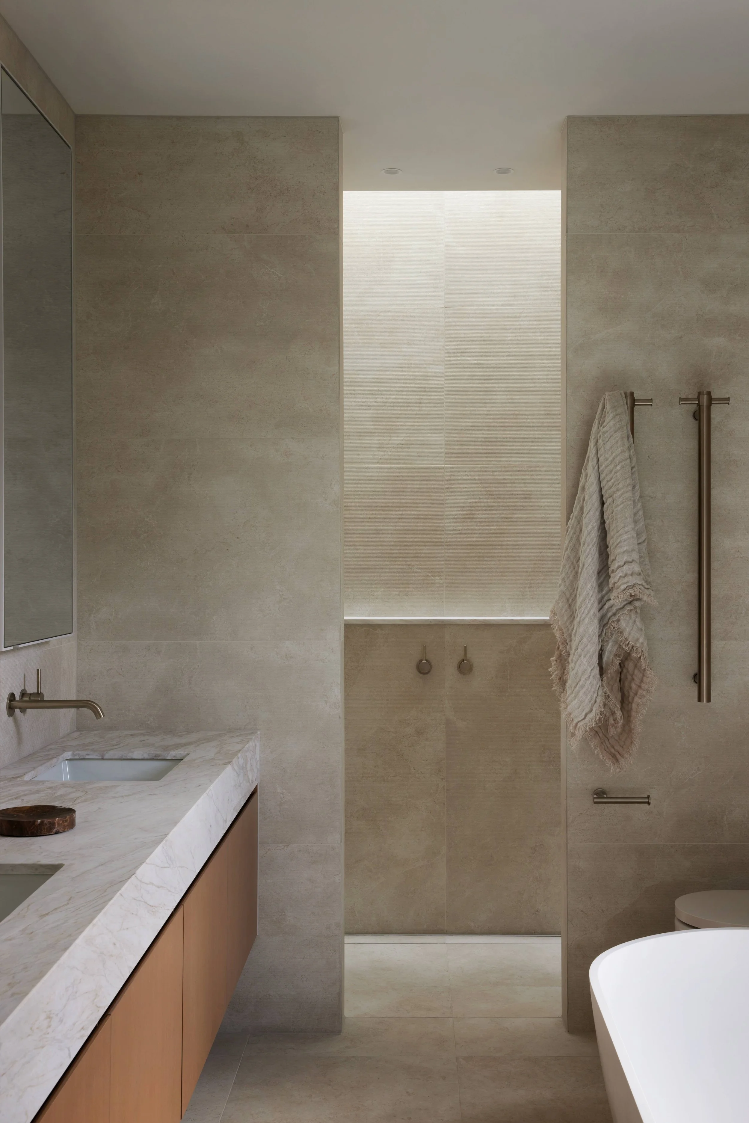 Modern bathroom featuring a marble countertop with two sinks, beige textured tiles on the walls, a frosted window with a ledge, towel hooks with a gray towel, and a partial view of a freestanding bathtub.