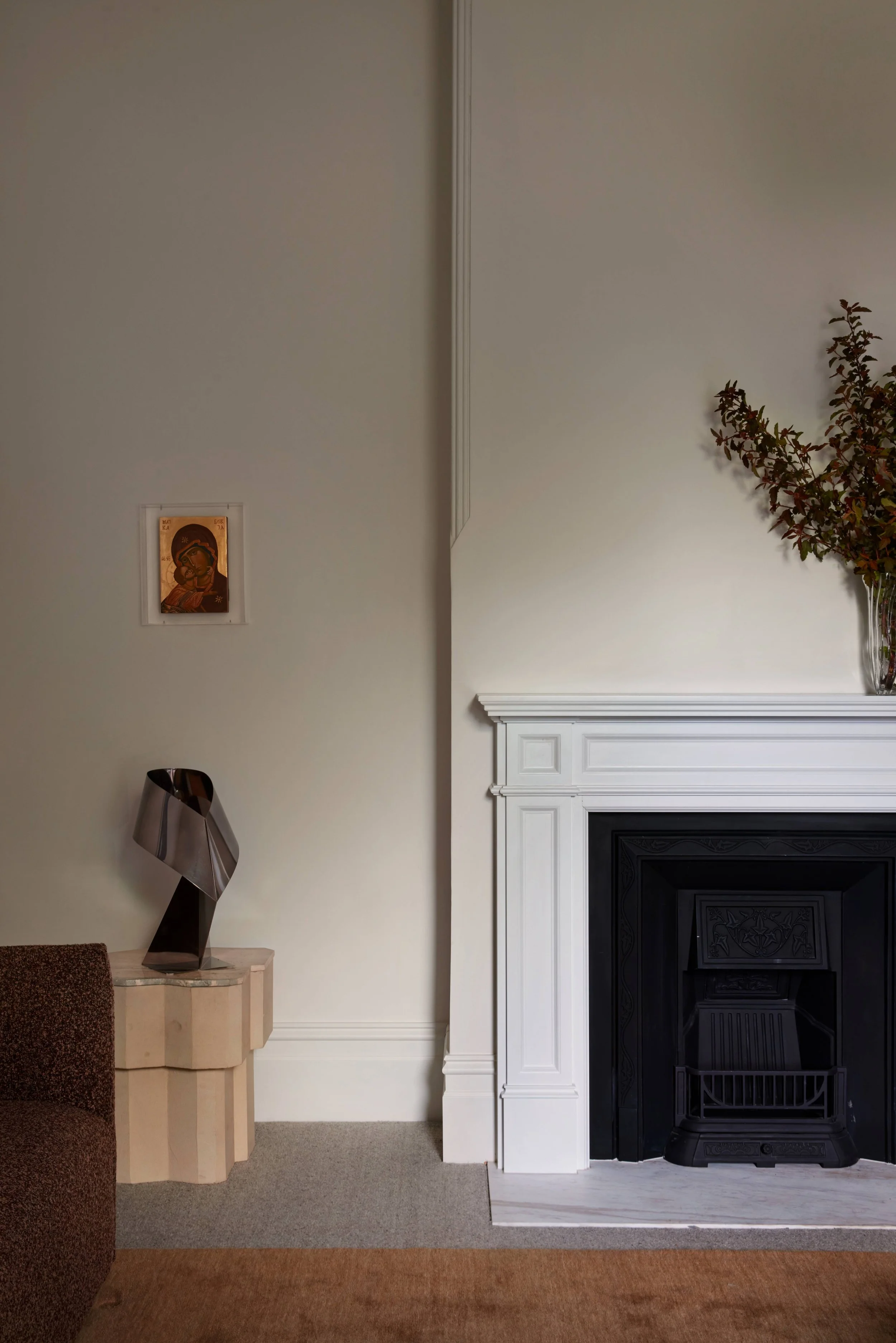 Living room with white built-in fireplace, a black insert, a potted plant on top of the fireplace, a small framed icon on the wall, a beige abstract sculpture on a ledge, and a brown textured chair partially visible on the left.