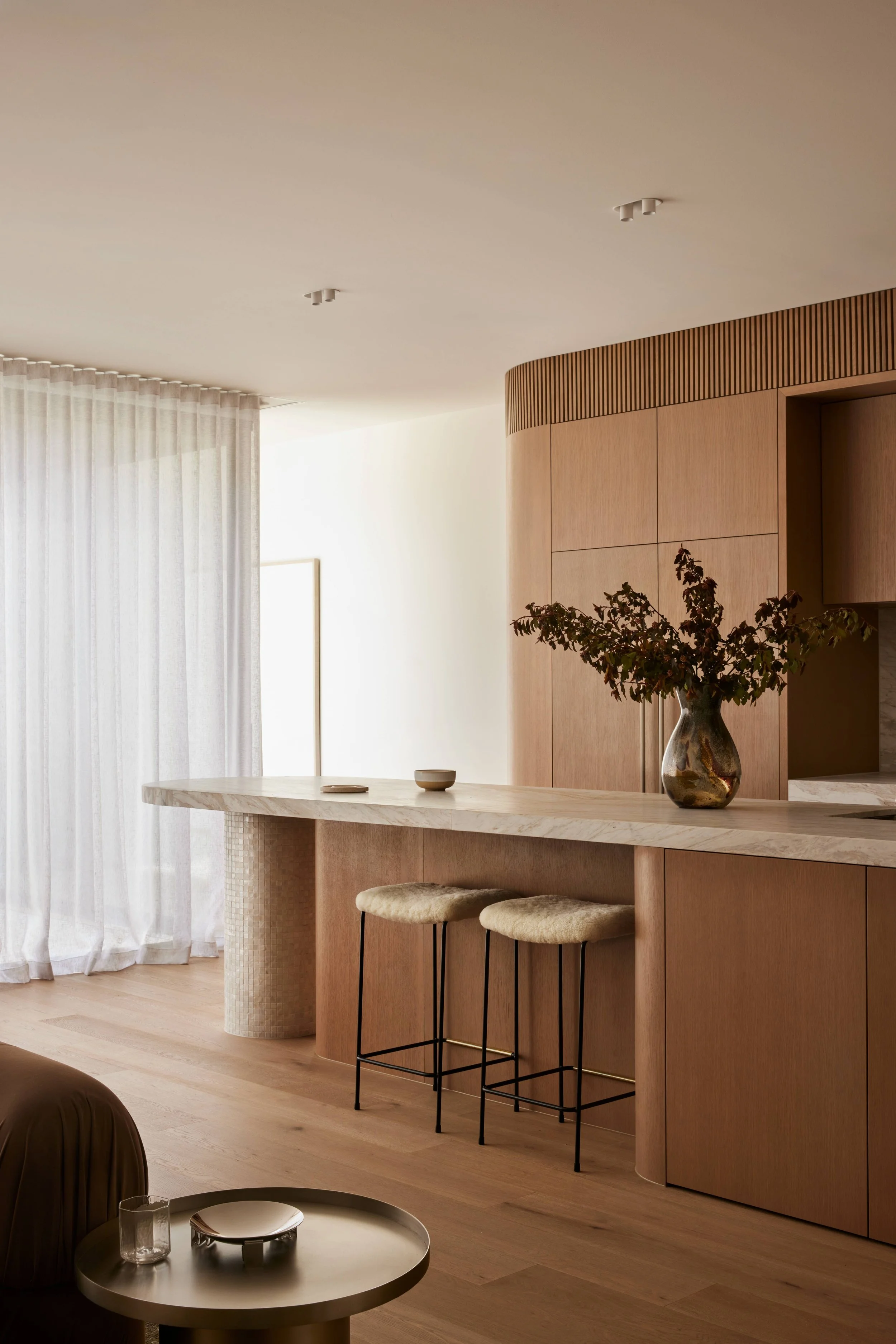 Interior of a modern, minimalist kitchen featuring a curved wooden cabinet, a beige marble countertop with two beige stools, a vase with greenery, and light wooden flooring with sheer curtains in the background.