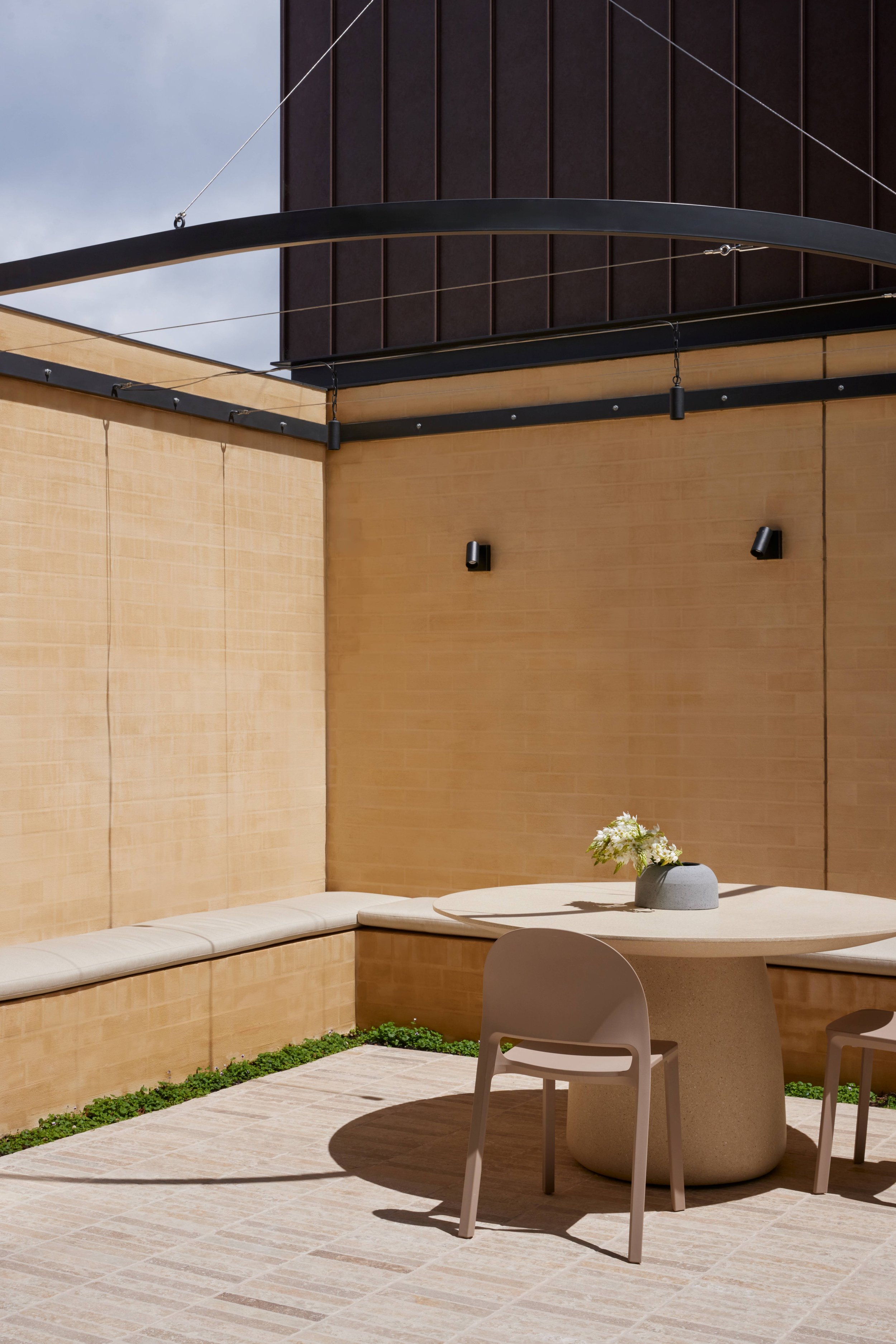 Outdoor seating area with a round table, two beige chairs, and a bench with cushions, set against tan walls with a potted flower centerpiece on the table and modern wall-mounted lights.