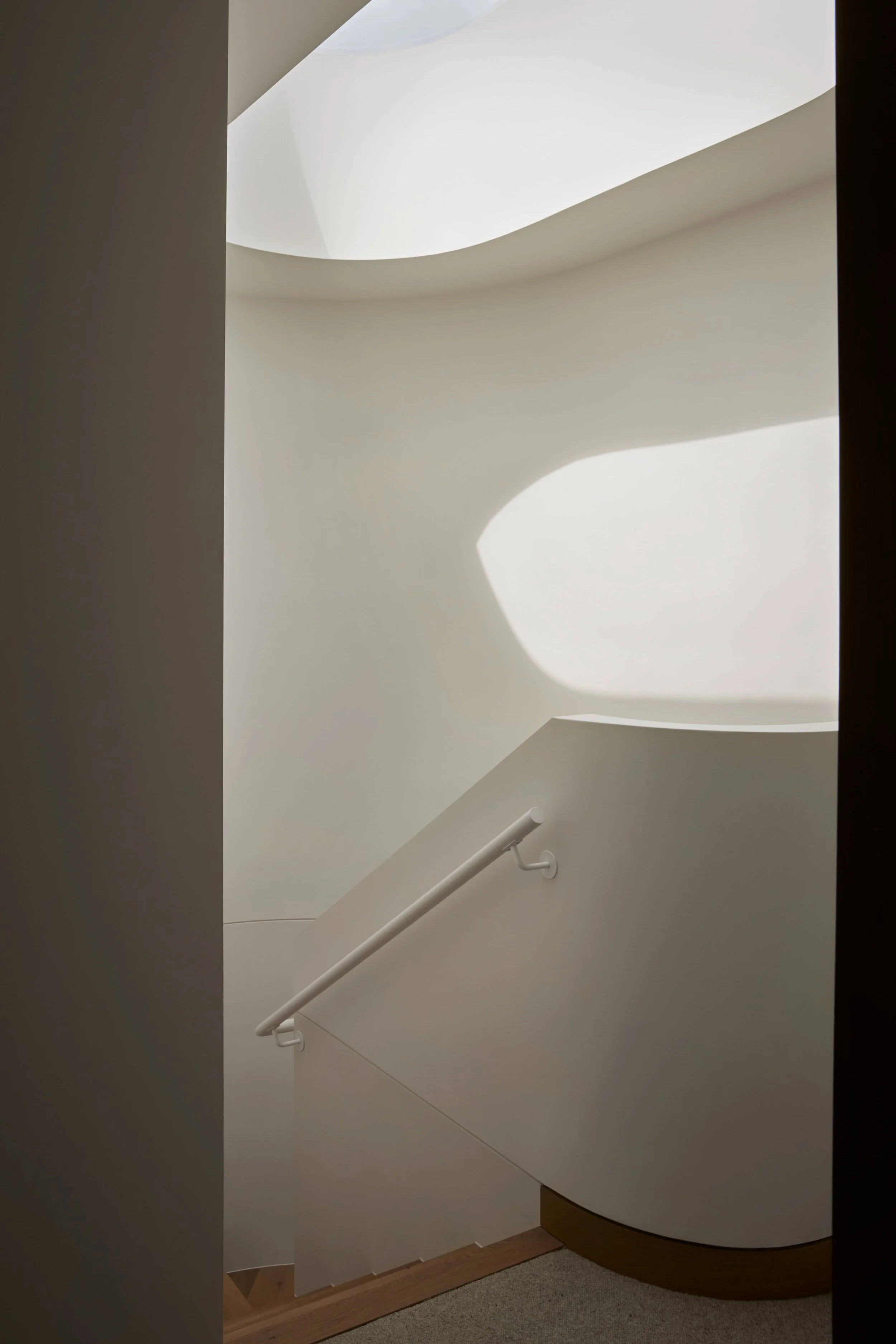 A modern staircase with smooth white walls and a ceiling with natural light coming through an opening, casting shadows.