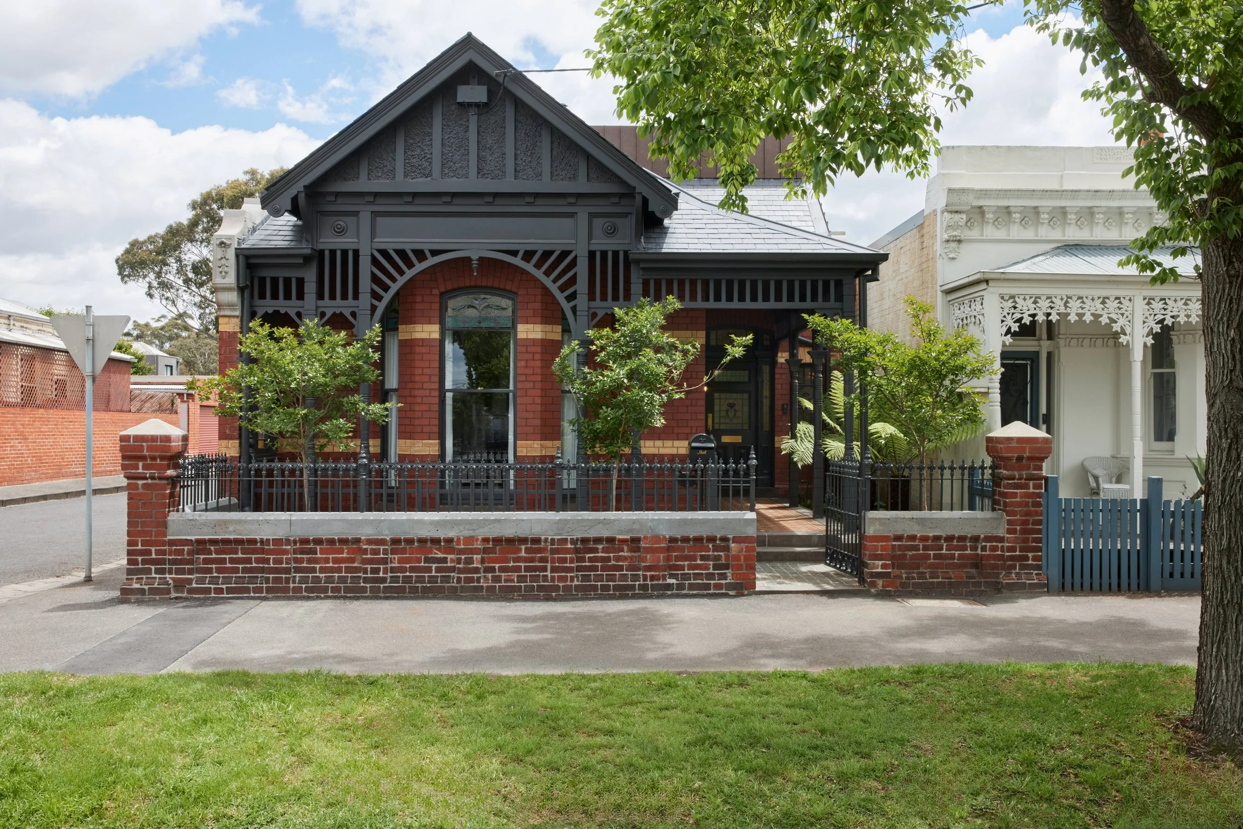 A house with a red brick facade, black decorative trim, and a front yard enclosed by a brick and metal fence, with trees and a sidewalk in front.
