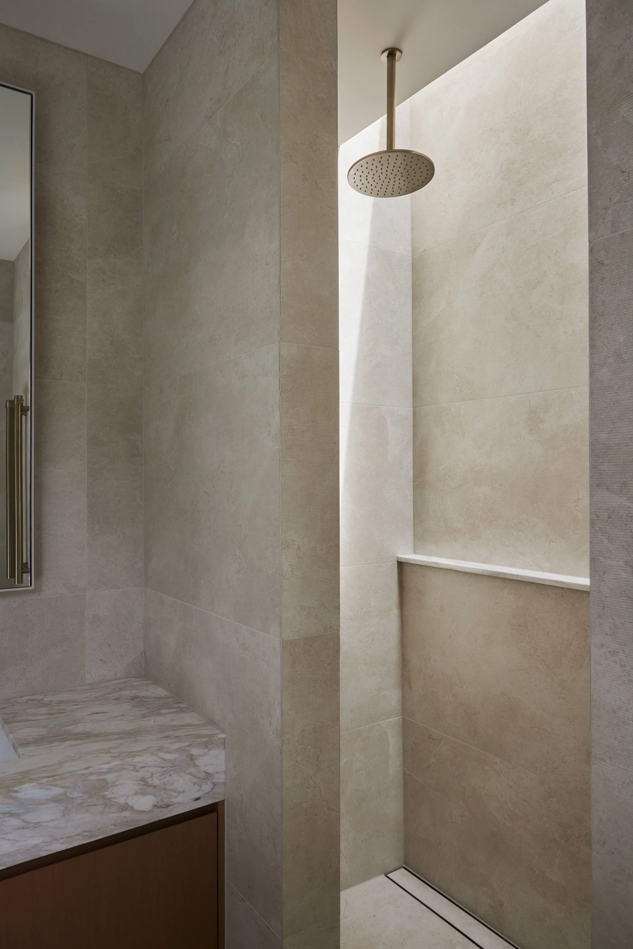 Modern bathroom shower area with beige tiled walls, a rainfall showerhead, and a built-in shelf. Part of a bathroom sink with a marble countertop and wooden cabinet is visible.