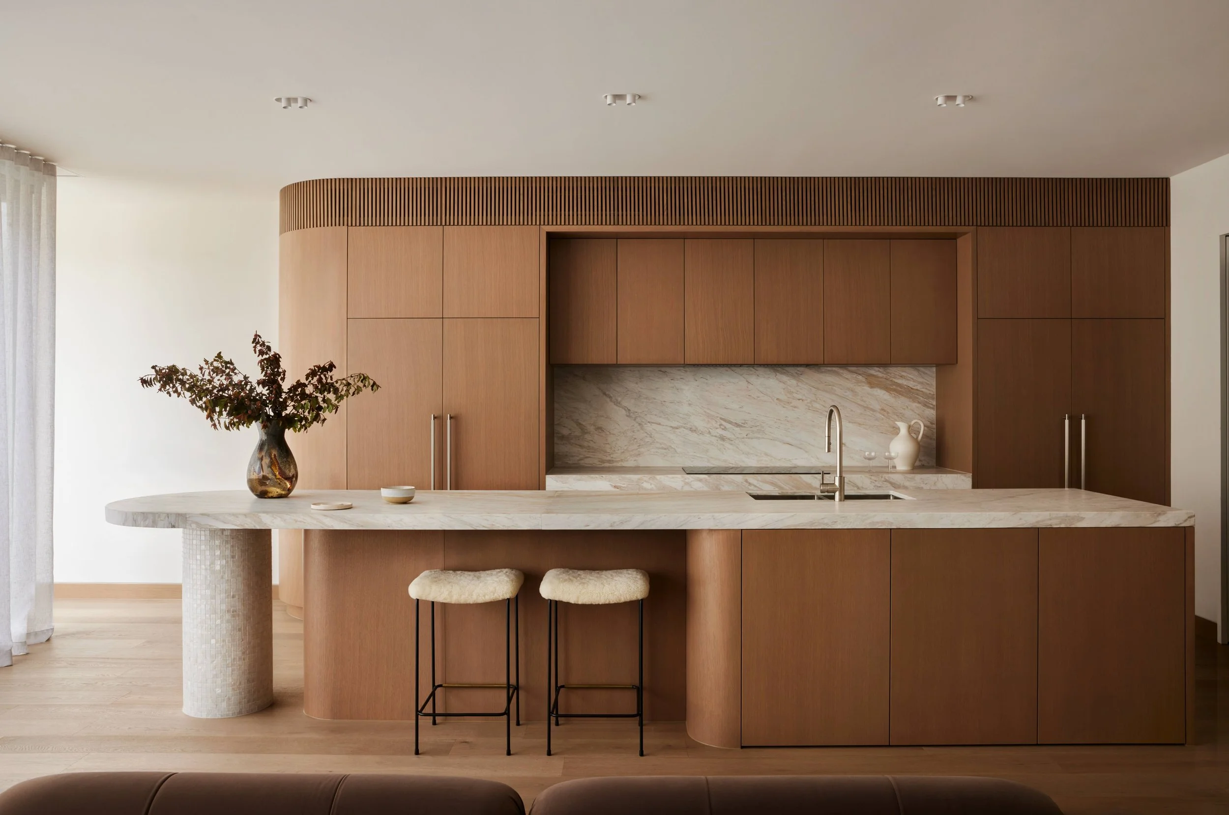 Modern kitchen with wooden cabinetry, marble countertops, a built-in sink, and two barstools with fur seats at the counter. A vase with greenery is on the left side of the island.