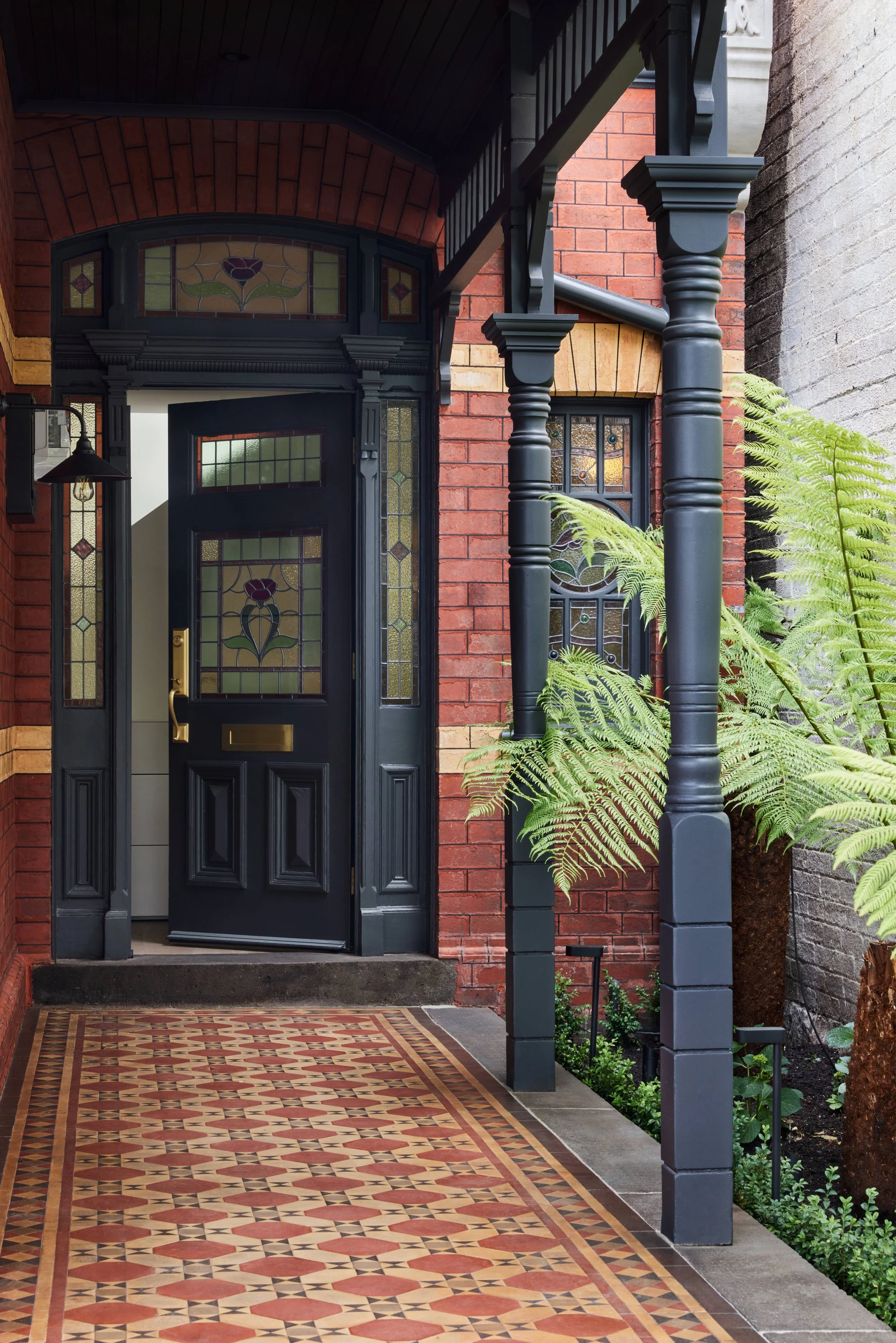 Front porch of a house with black door, stained glass windows, brick walls, fern plants, and patterned tile flooring.