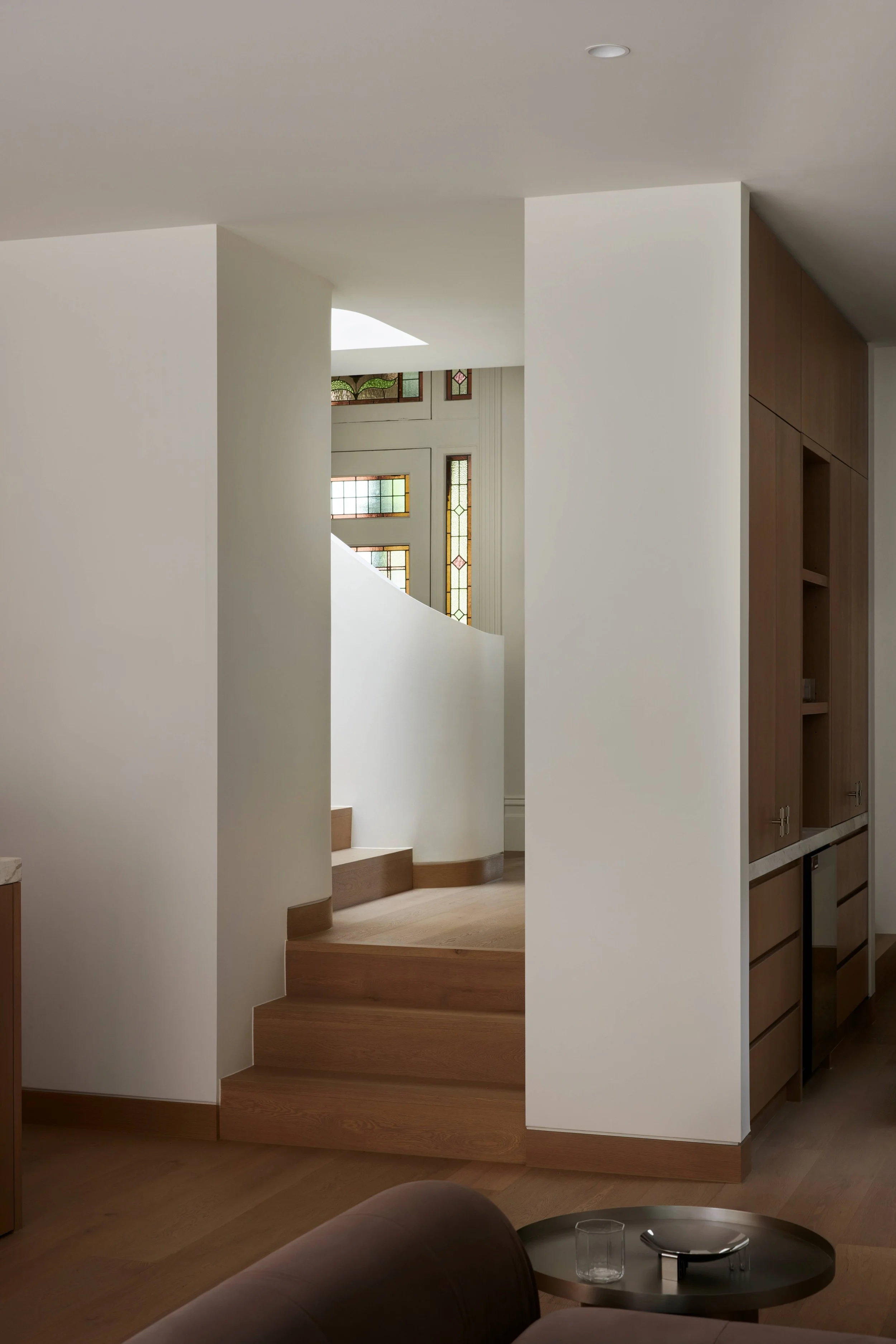 Interior view of a modern house featuring a small staircase with wooden steps, white walls, a built-in wooden cabinet, and decorative stained glass windows.