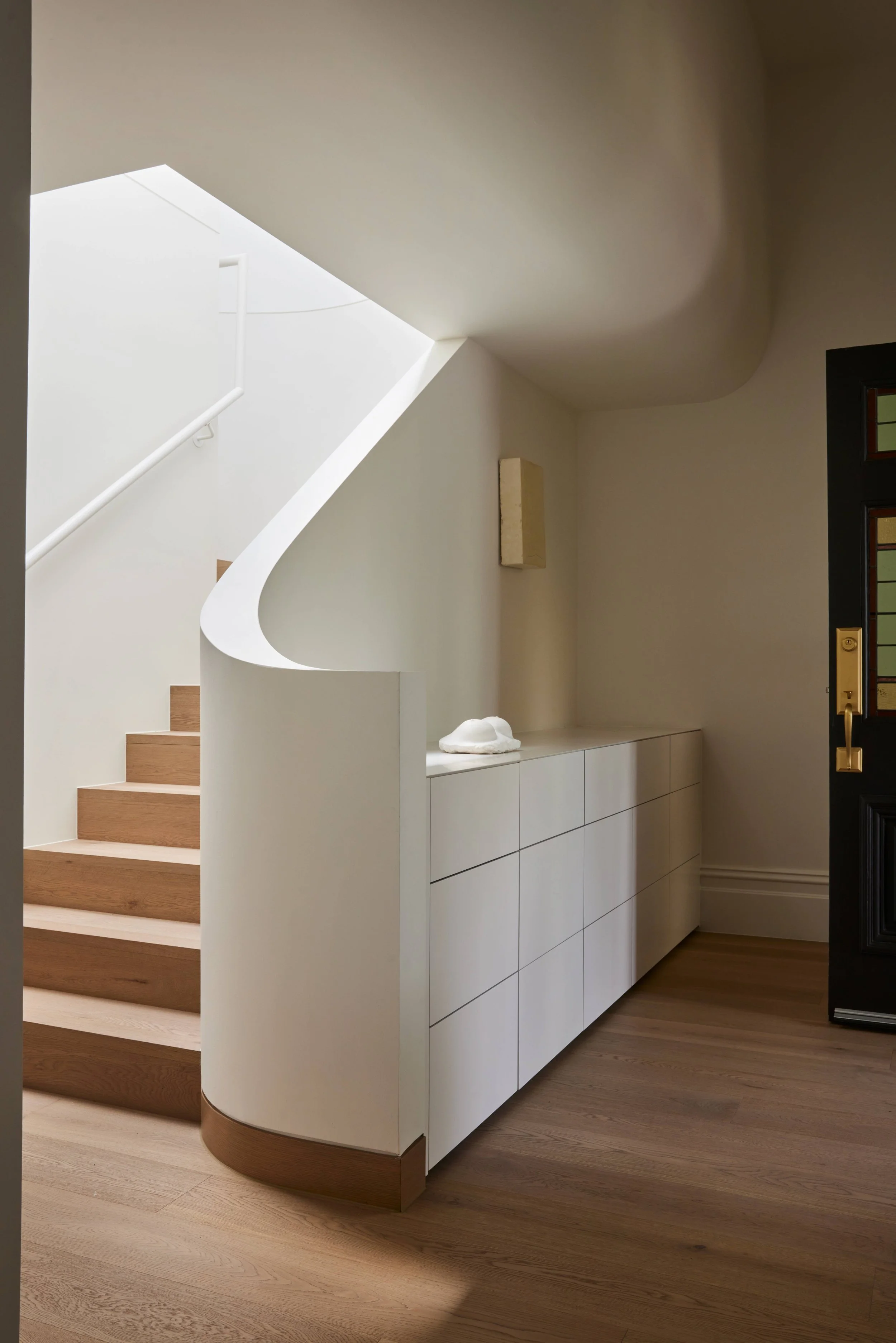 Interior view of a modern entryway with wooden stairs, white curved wall, white cabinet, and black door.