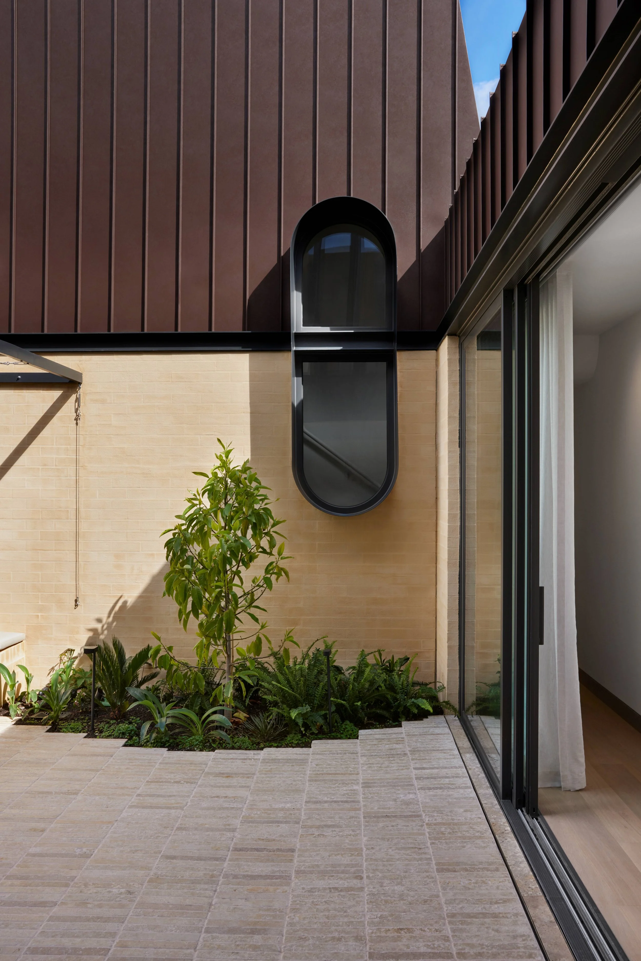 Modern house exterior with beige brick wall, brown metal paneling, a curved black-framed window, and a sliding glass door leading to an indoor space with a curtain. A small garden with green plants and a young tree is situated on a tiled patio.