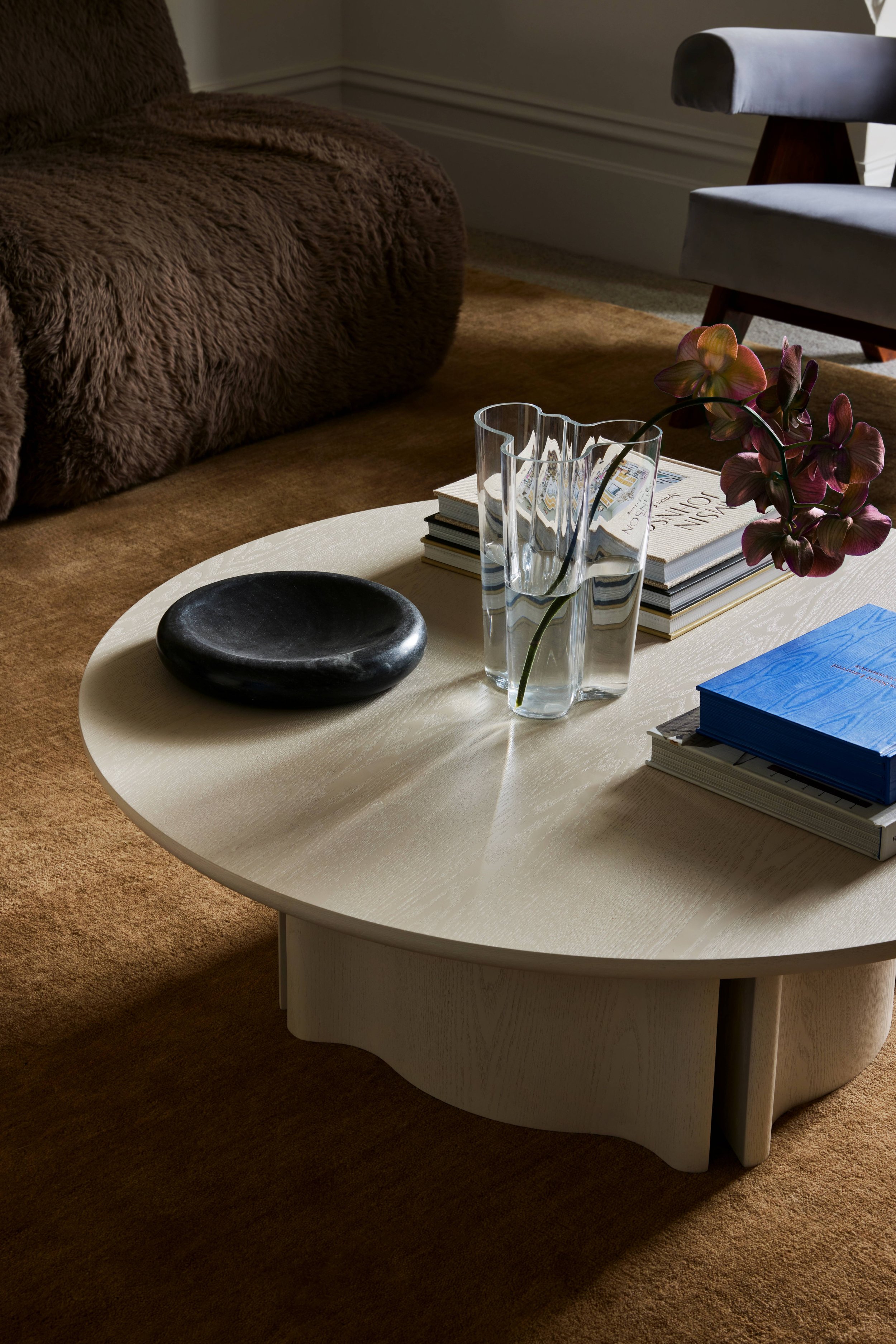 A round, off-white wooden coffee table with a black stone dish, a tall glass vase with a pink orchid, and stacked books, in a cozy living room with a brown fuzzy sofa and a gray armchair.