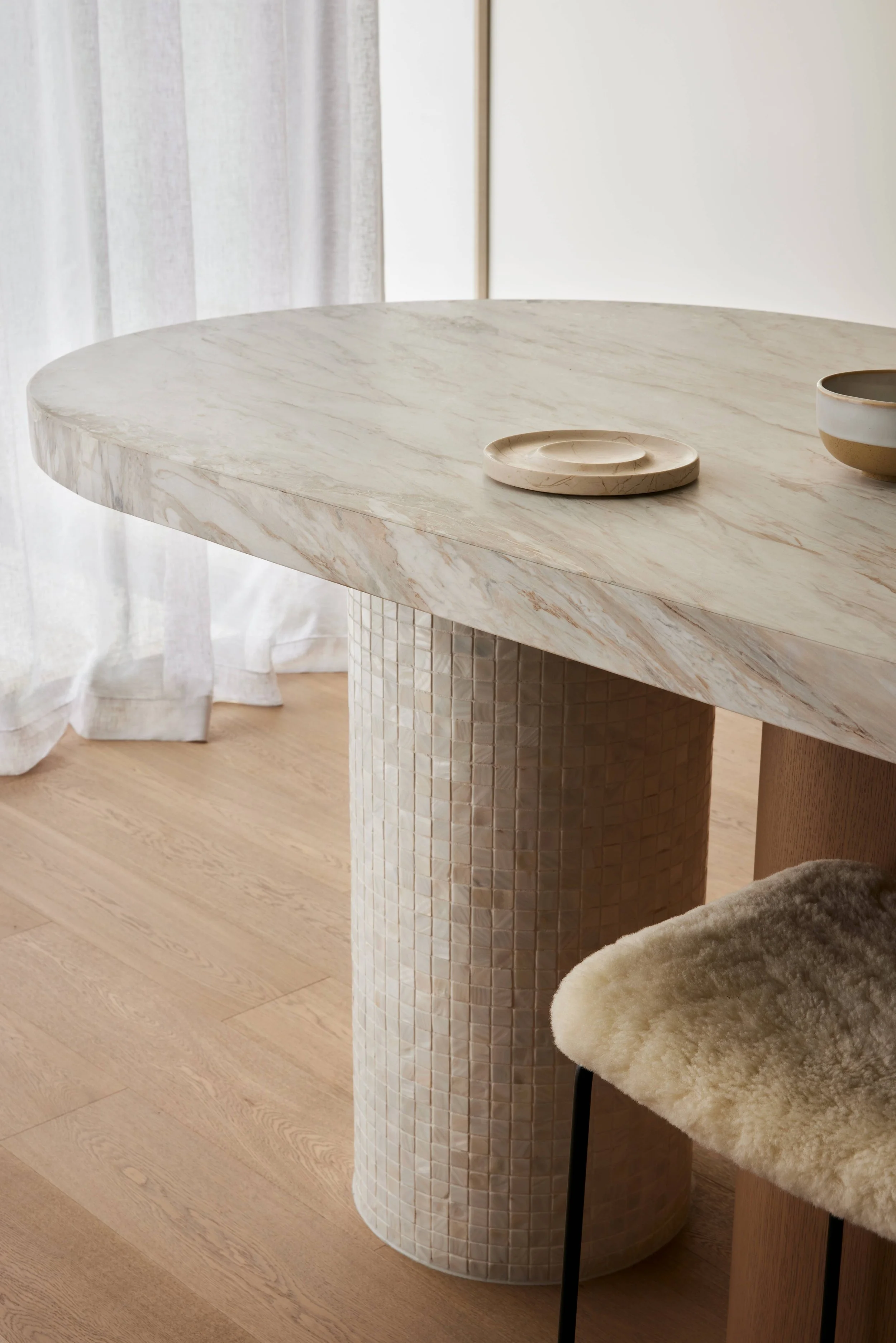 A round marble dining table with a textured cylindrical base, set with small decorative plates and bowls, next to a cream-colored fur-lined chair in a bright, airy room with light wood flooring and semi-transparent curtains.