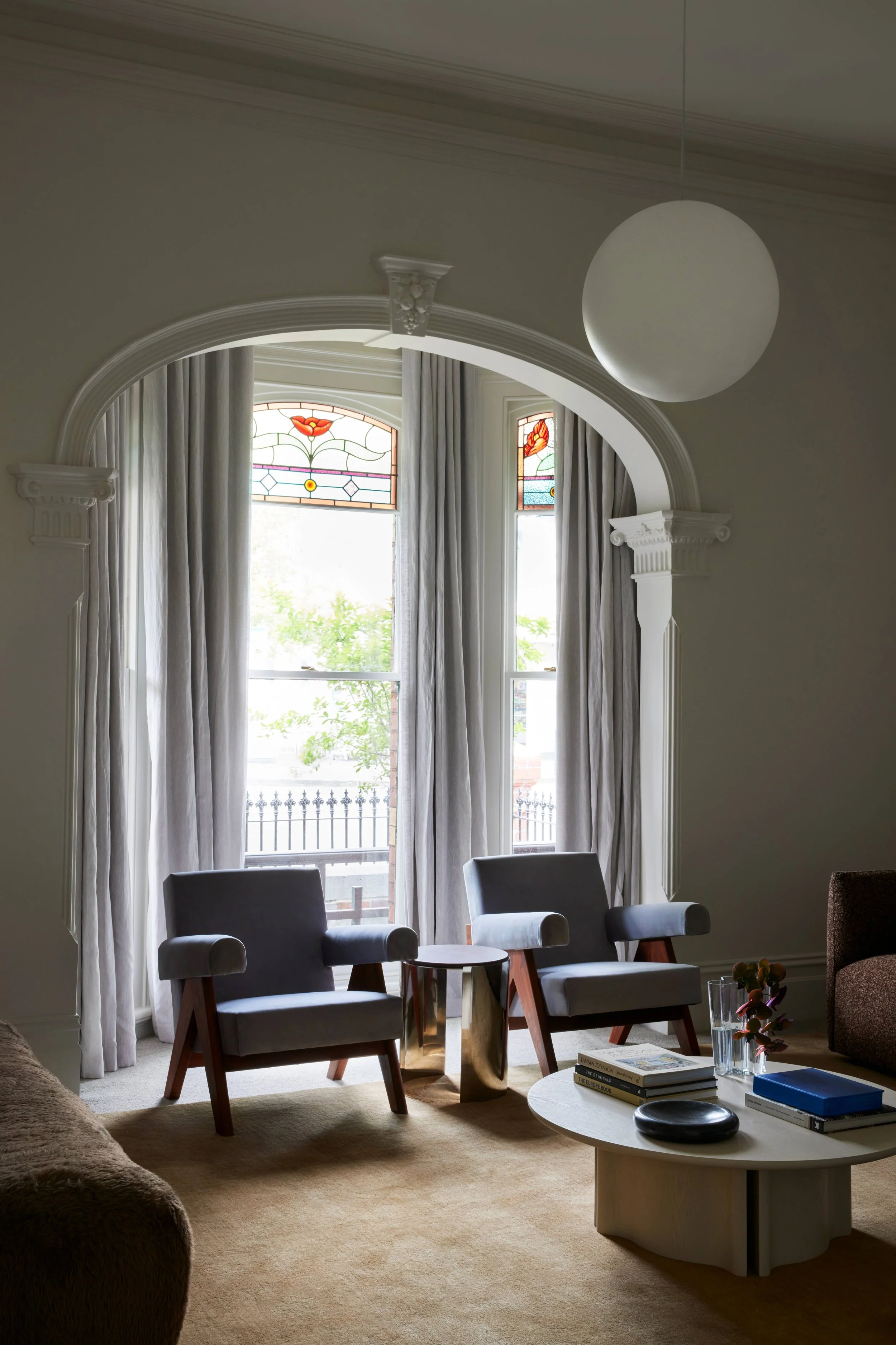 Living room with two mid-century modern armchairs, a small round side table, a coffee table with books and decorative objects, large windows with stained glass accents, and cream-colored curtains.