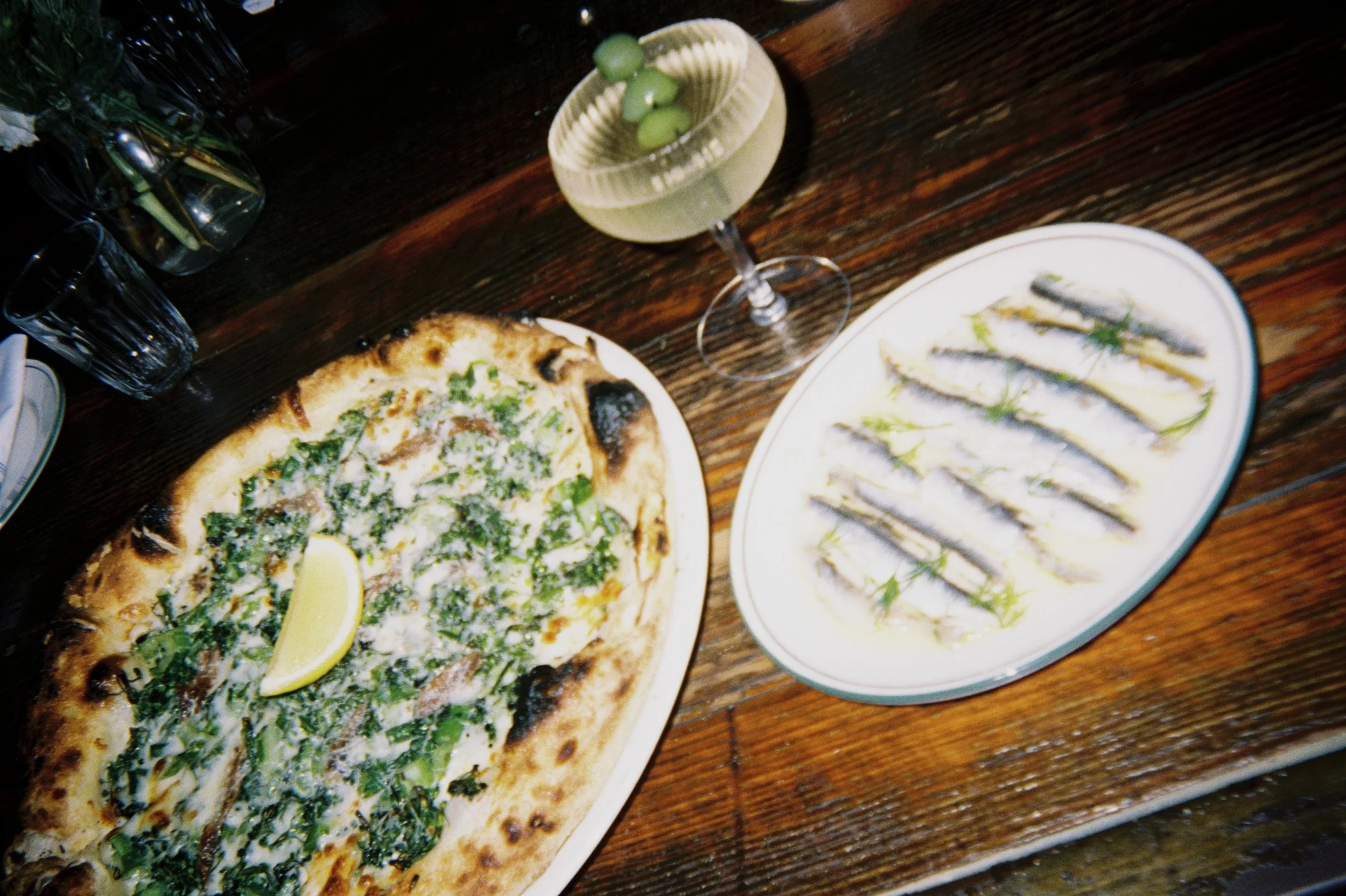 A pizza with green herbs and lemon wedges, a plate of sushi rolls, and a cocktail with green olives on a wooden table.