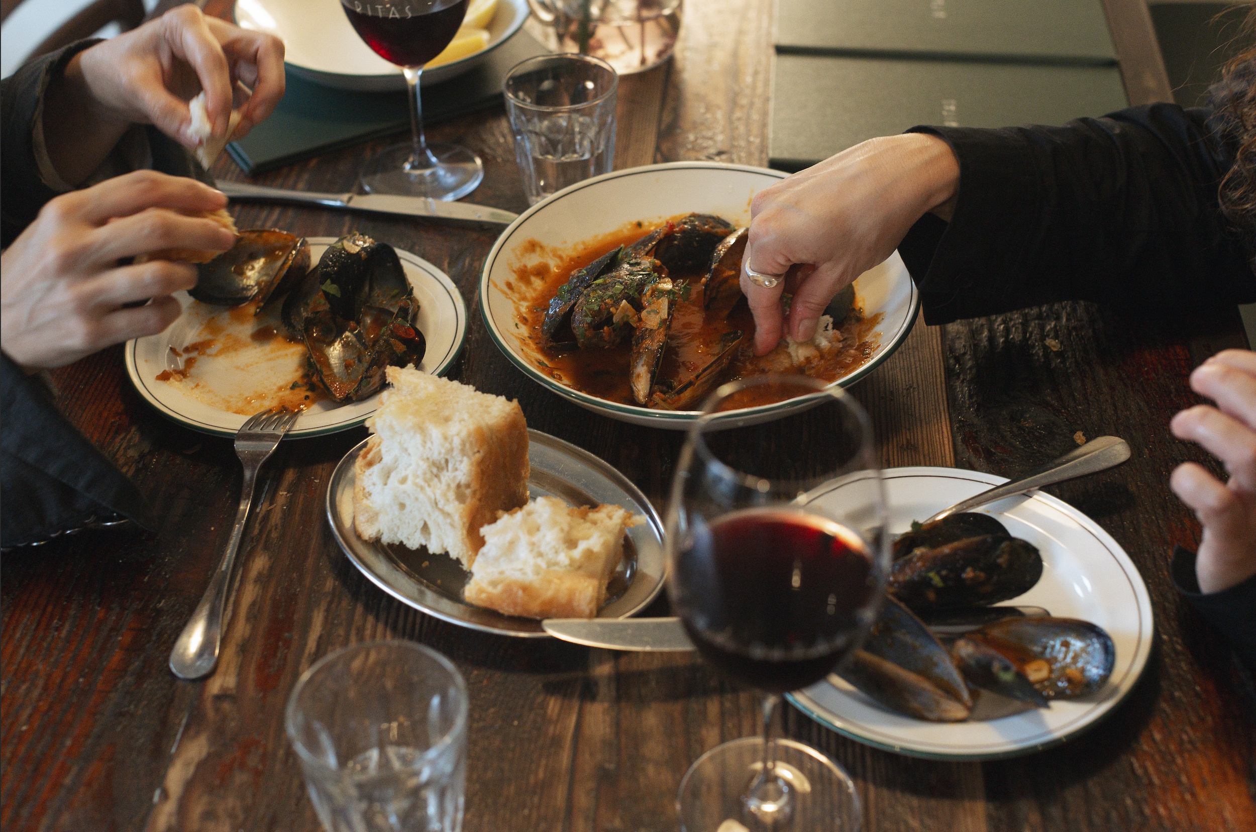 People eating a meal of fish with sauce, bread, and drinks including red wine and water on a wooden table.