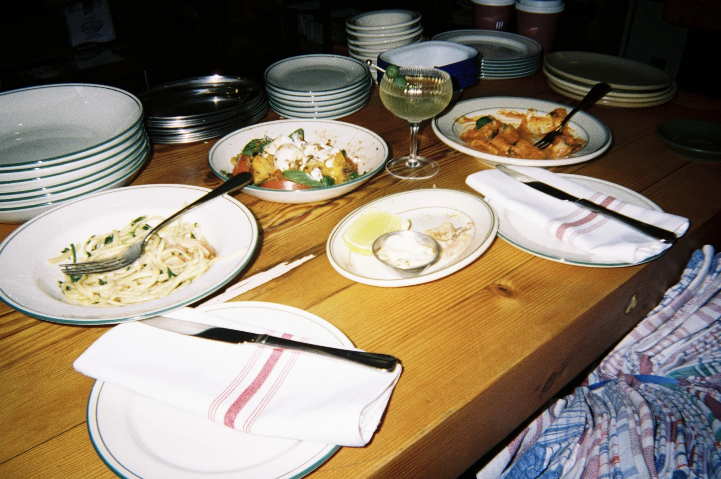 A wooden table set with plates of pasta, a vegetable dish, a lemon wedge, a glass of white wine, and stacks of dishes and bowls in the background.