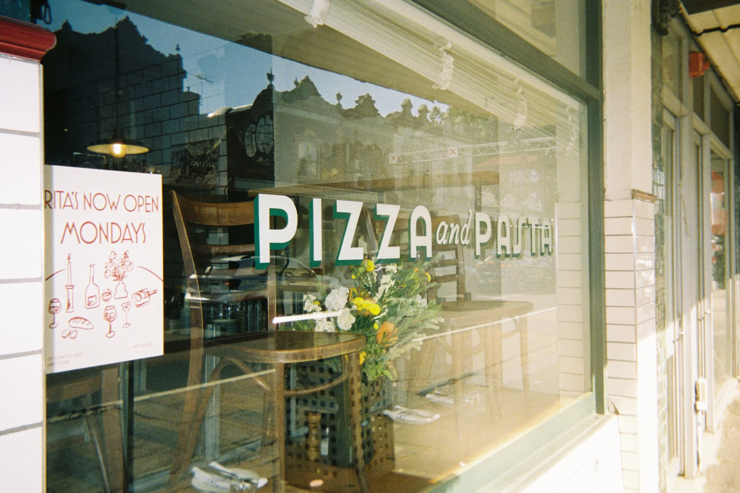 A storefront window advertisement for a pizza and pasta restaurant, with a sign reading 'RITA'S NOW OPEN MONDAYS', visible chairs, flowers, and reflections of buildings and trees.