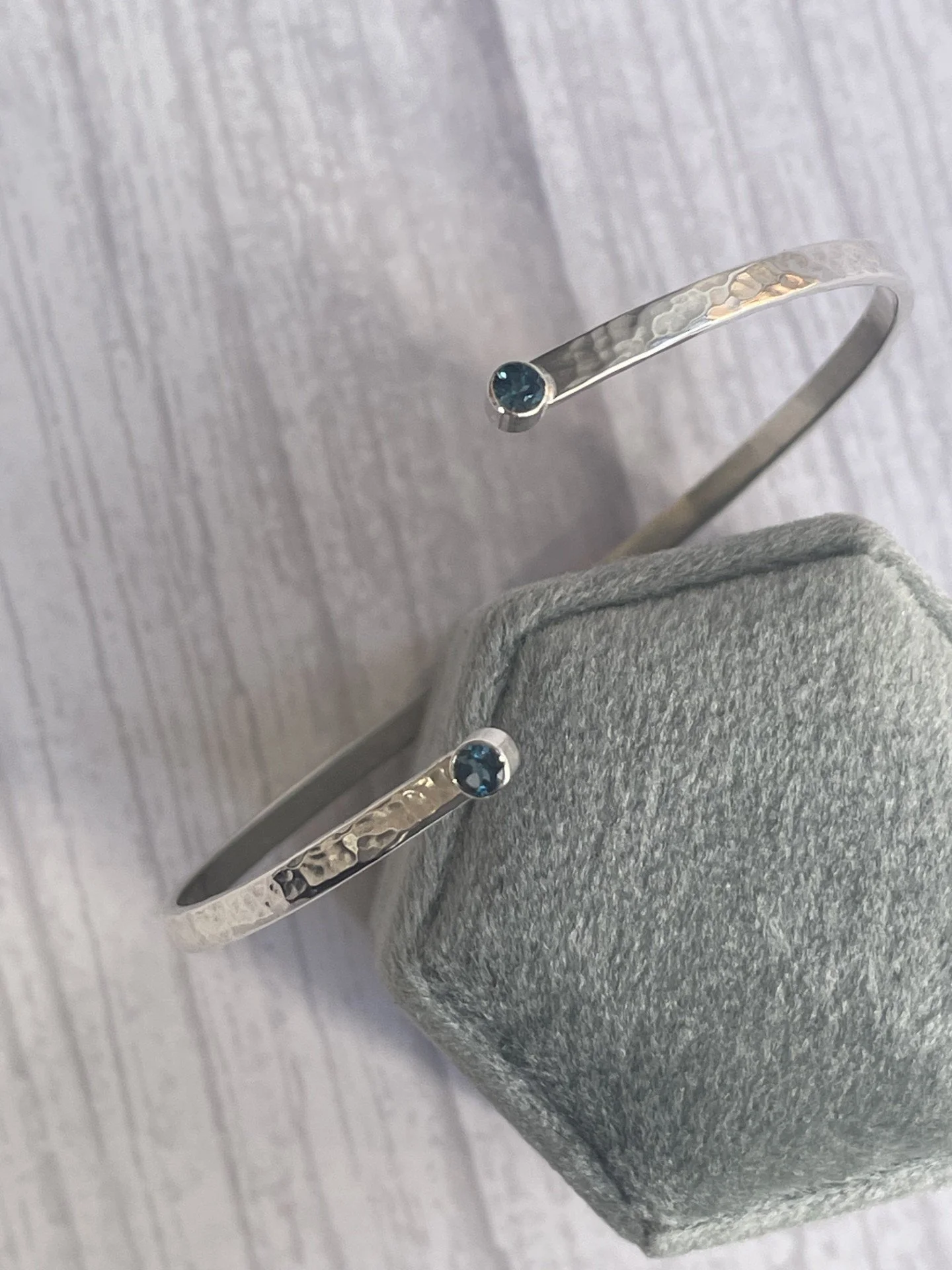 Silver bracelets with a small round blue gemstone each, placed on a gray textured jewelry holder.