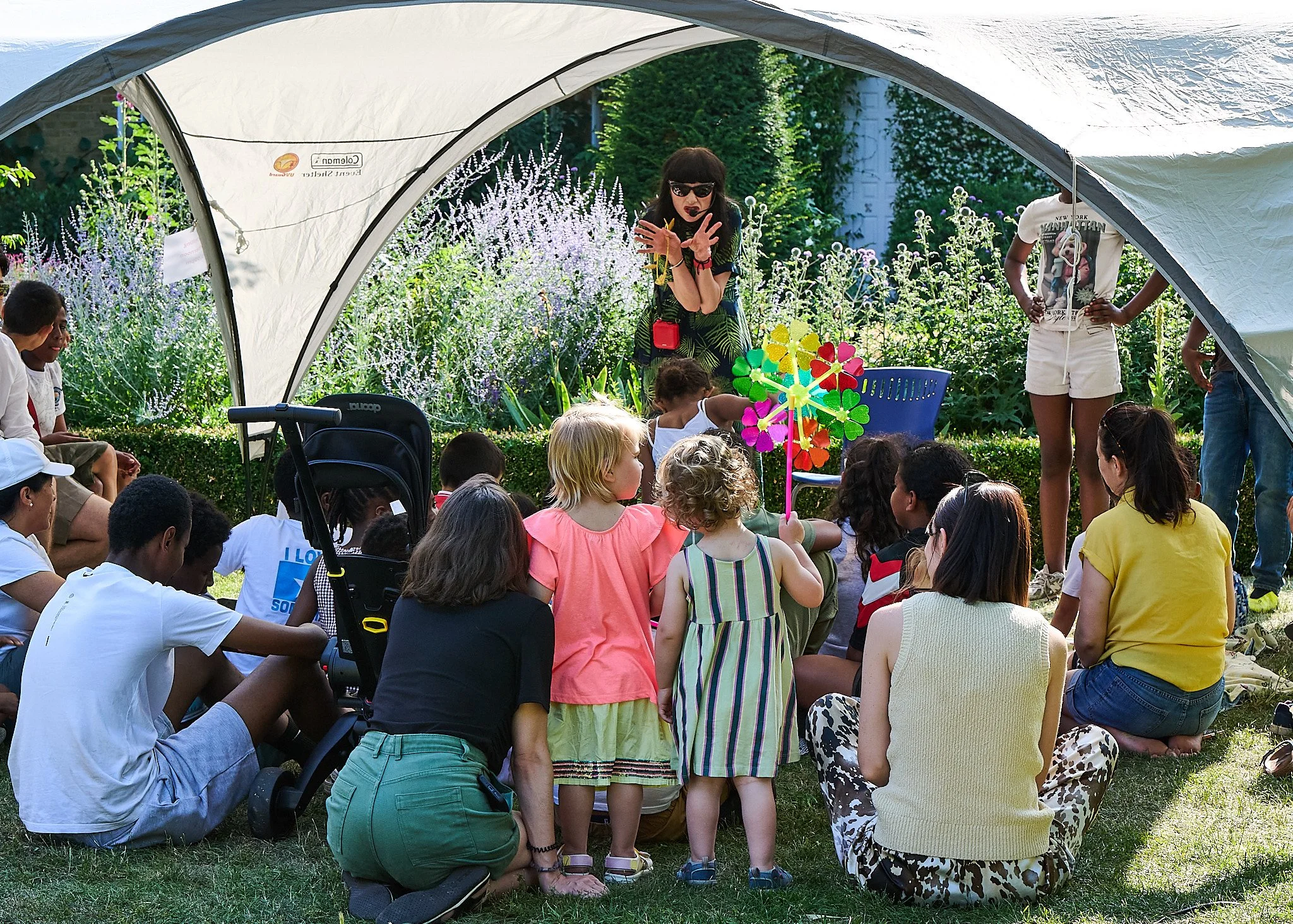 Group of children gathered outdoors under a white canopy listening to a woman with sunglasses and a headset. The woman is standing in front of colourful pinwheels, surrounded by lush greenery and flowers. She is performing a story using drama.